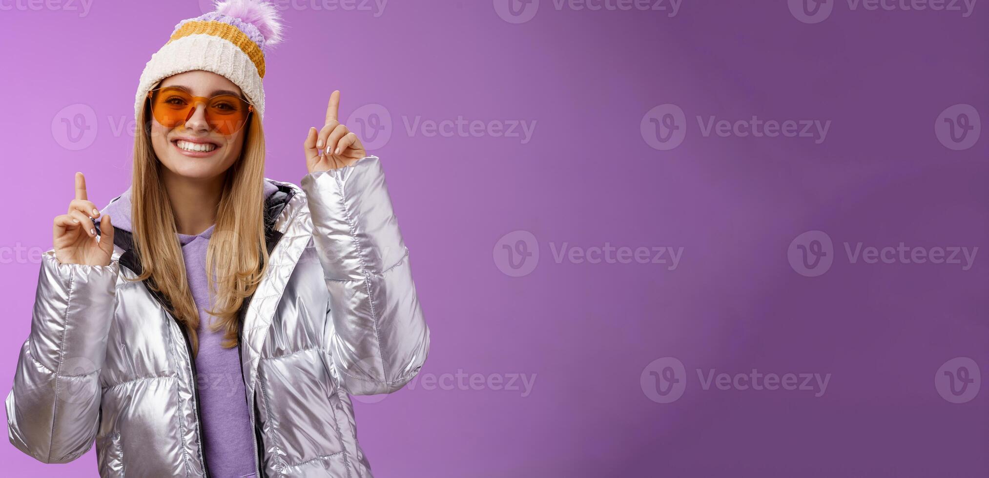 Joyful energized entertained cute blond woman having fun enjoy vacation snowy mountain trip wearing sunglasses silver jacket winter hat dancing pointing up amused standing purple background photo