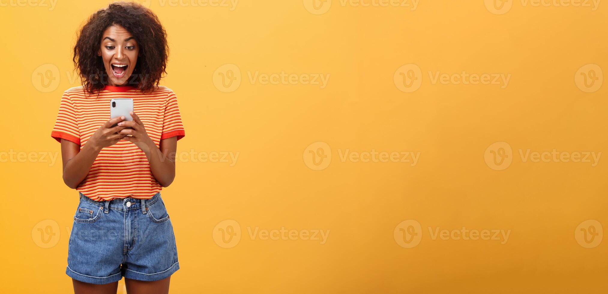 Girl expressing excitement and joy receiving awesome invitation via messages yelling from delight and happiness looking at smartphone screen impressed and thrilled over orange background photo