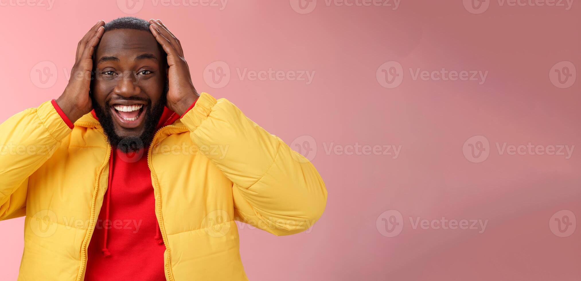 Happy surprised pleased young african-american 25s man young jacket red hoodie smiling amused, astonished smiling receiving awesome gift holding hands head thrilled, standing pink background photo