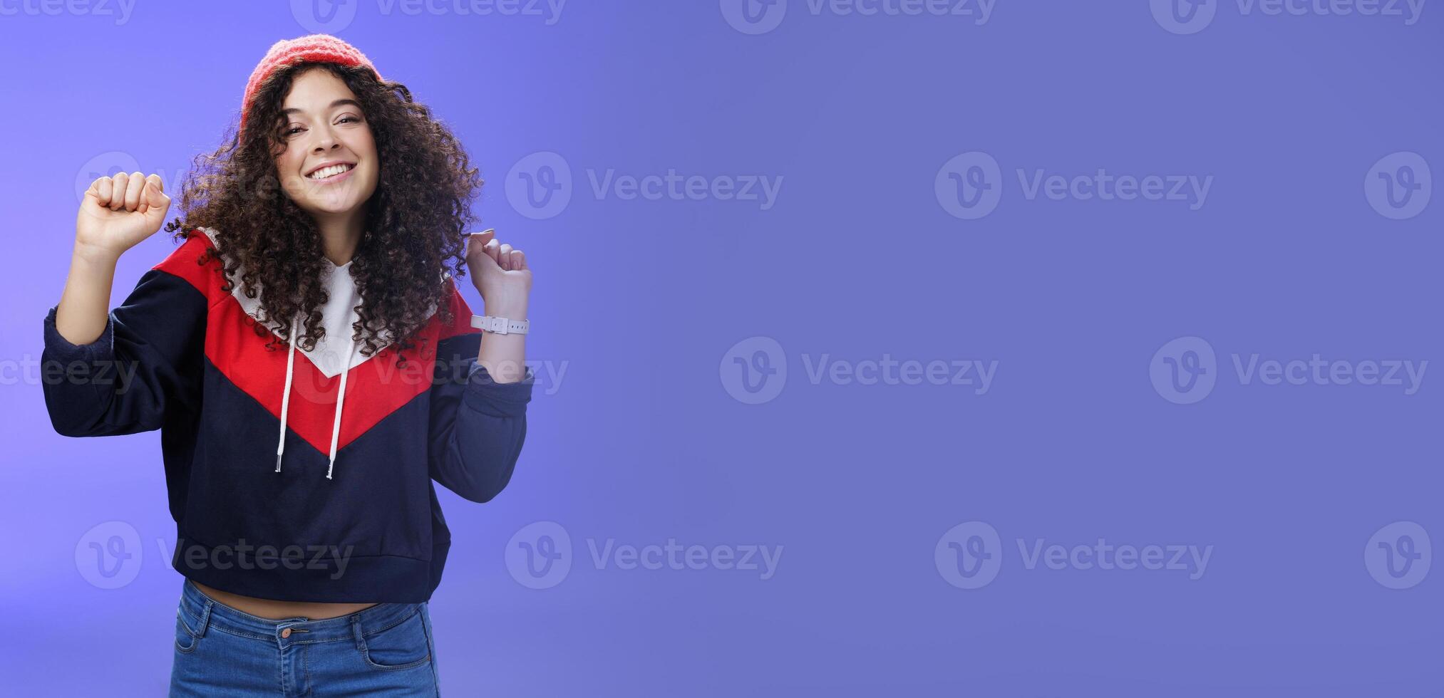 Charming carefree young 20s girl with curly haircut in warm beanie dancing and having fun joyfully over blue wall raising hands relaxed, chill smiling broadly at camera liking perfect chilly weather photo