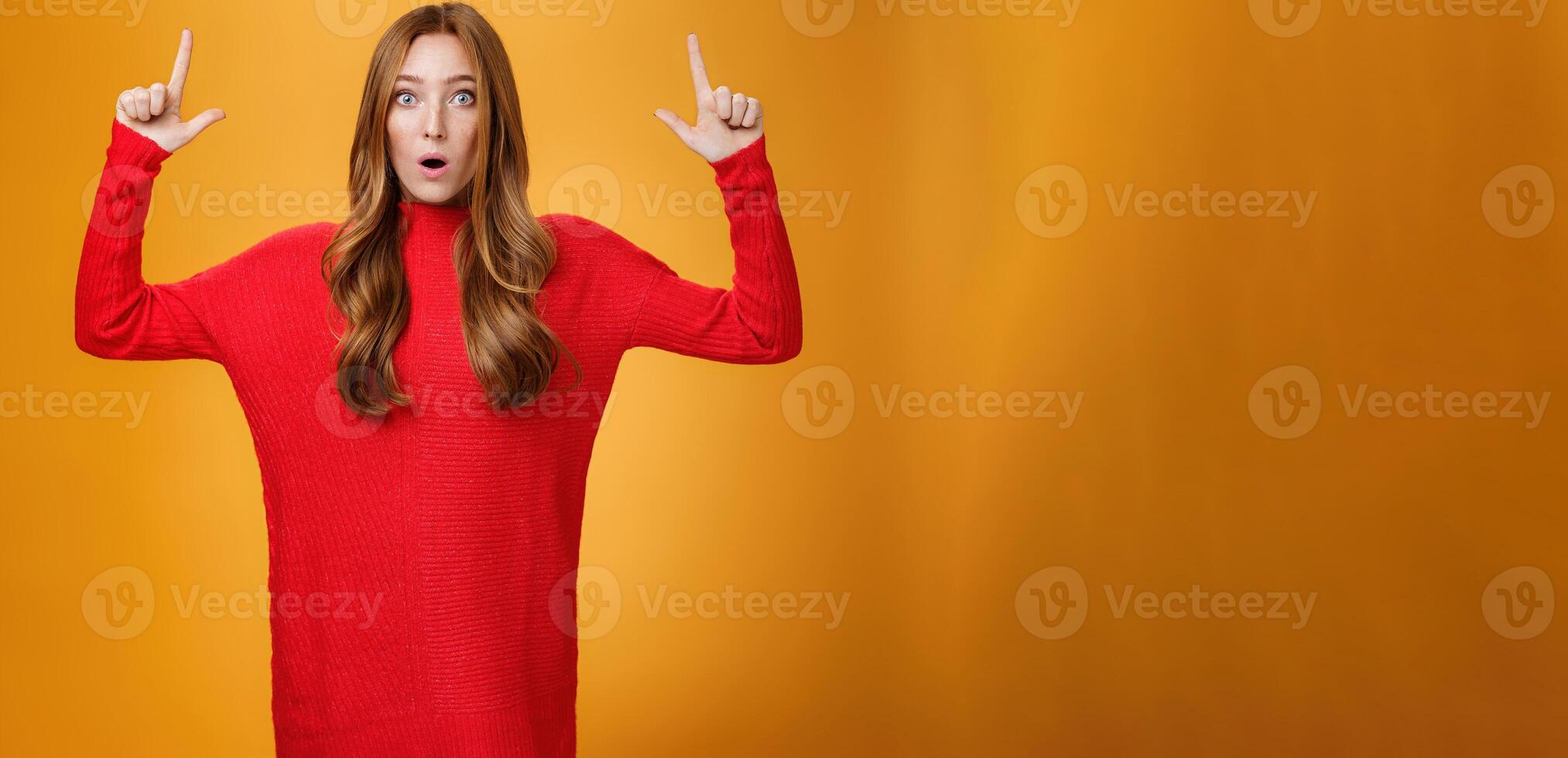Portrait of attractive and stylish redhead 25s female in knitted red dress gasping from amazement open mouth questioned and surprised as pointing up at astonishing promotion over orange wall photo