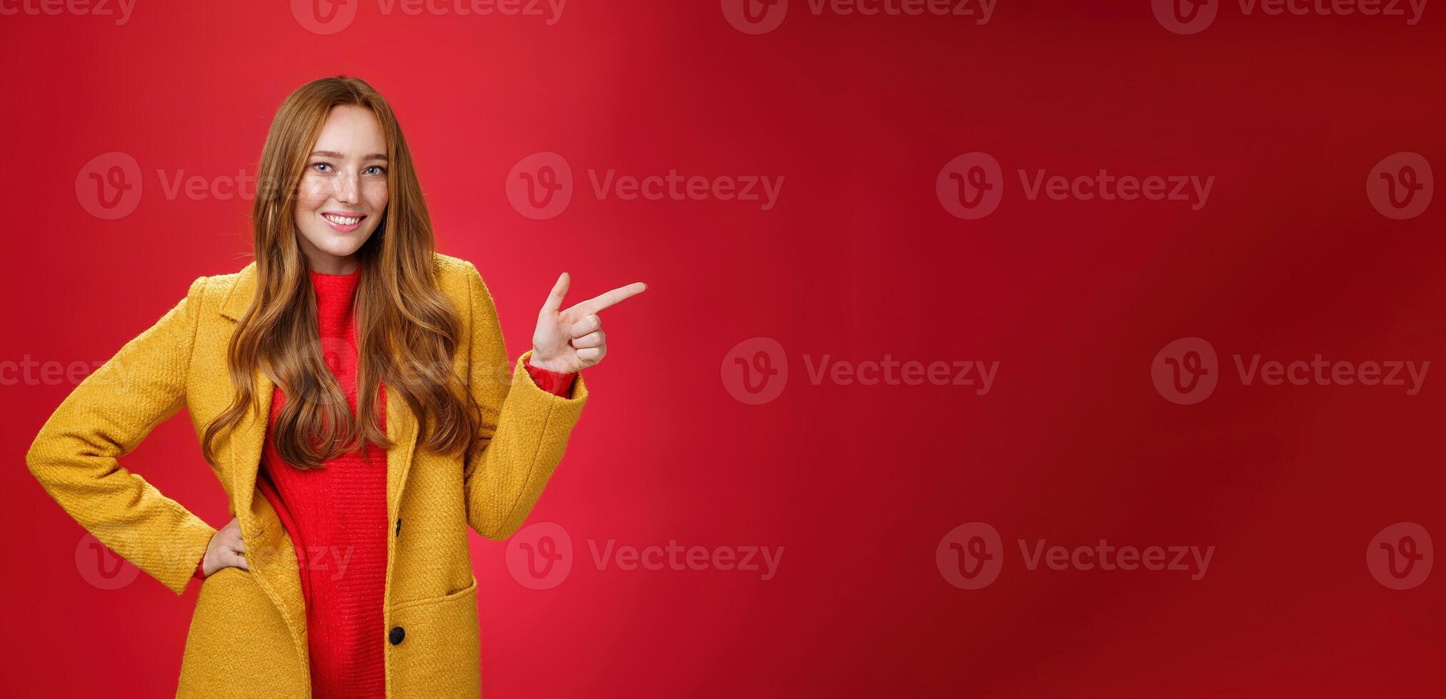 Stylish and self-assured attractive redhead woman with freckles in yellow outdoor coat pointing right with finger pistol and smiling joyfully as camera as showing cool promotion over red wall photo