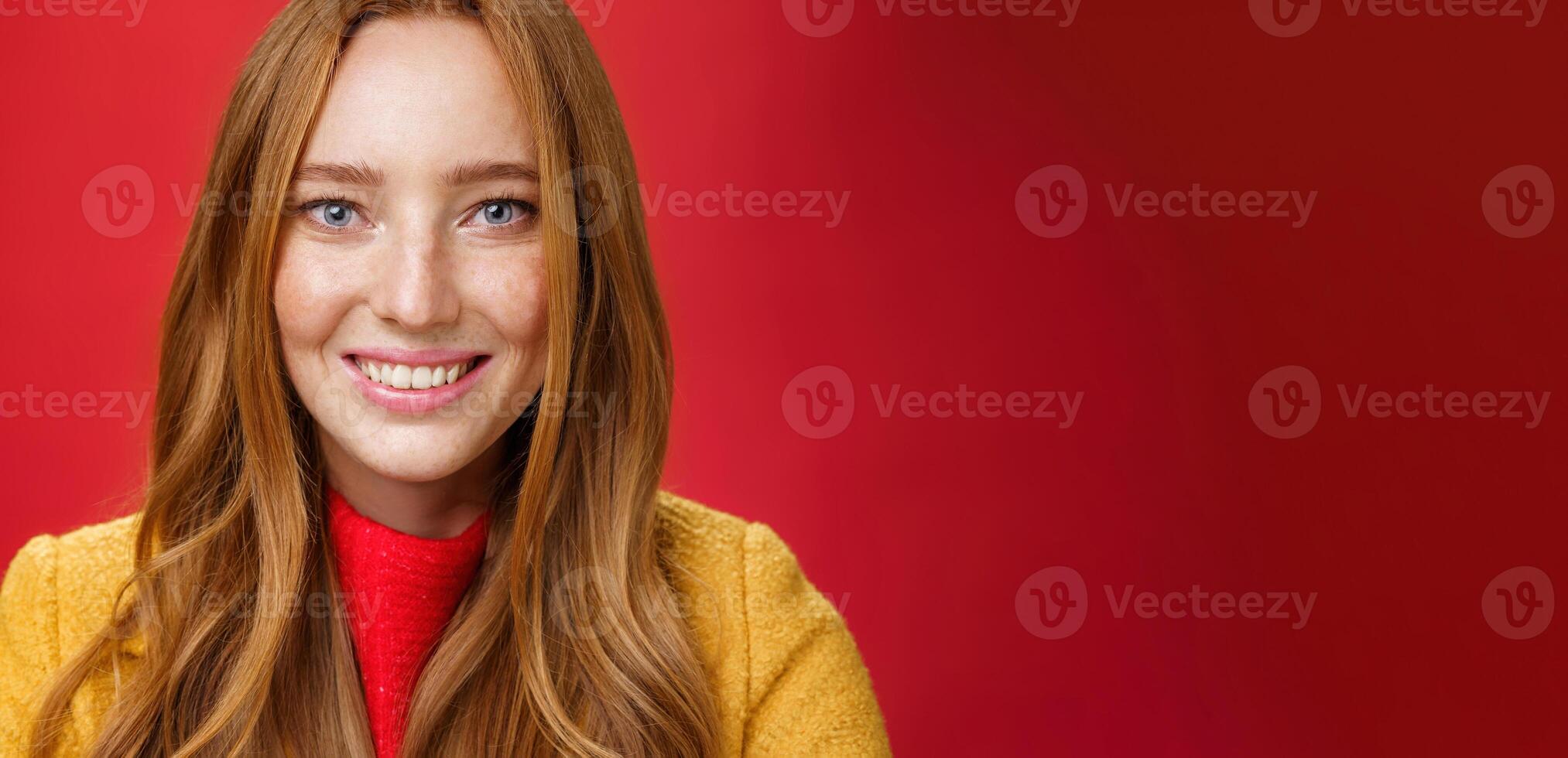 Headshot of hopeful and happy young redhead 20s girlfriend with cute freckles and blue eyes smiling broadly with faithful excited expression having high hopes, posing over red background photo
