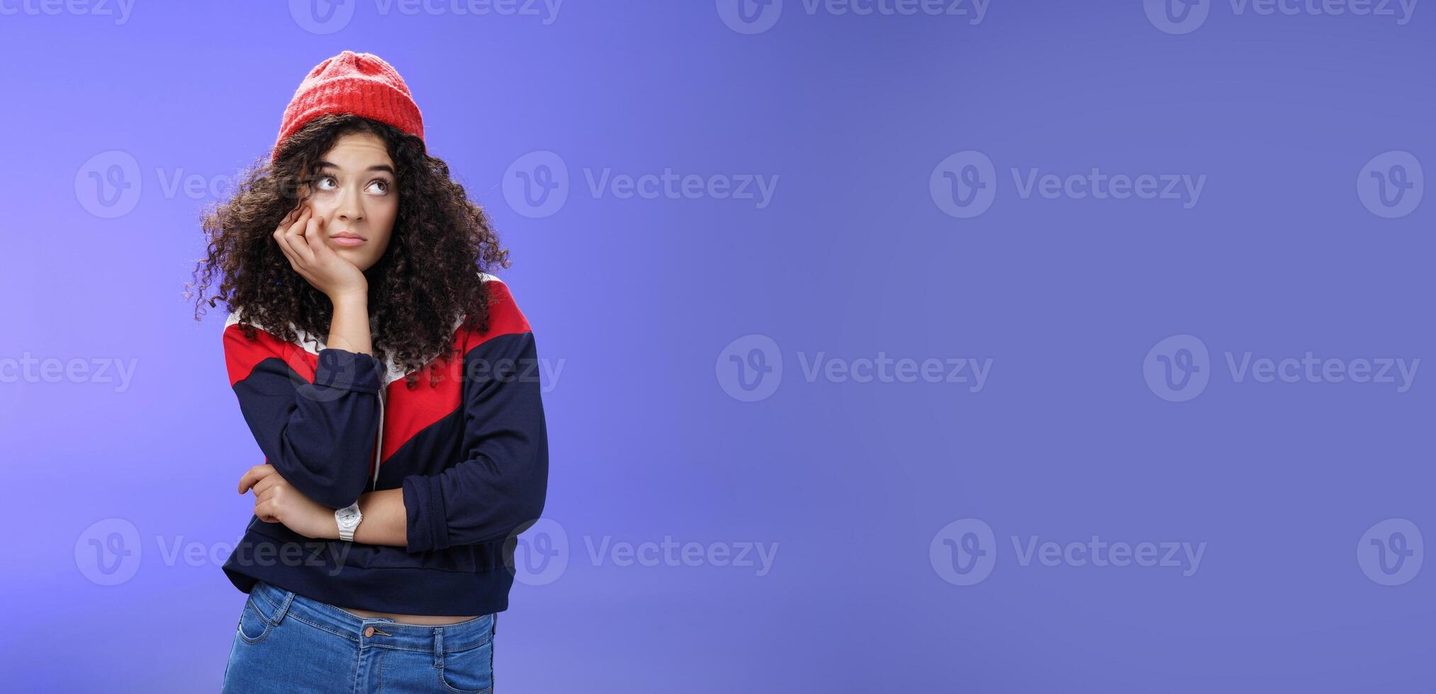 Portrait of frustrated and bored lonely girl with curly hair in winter hat holding hand across chest leaning head on palm, looking silly and gloomy at upper right corner, thinking feeling boredom photo
