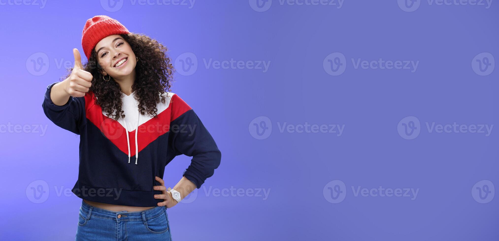 You doing great, like. Portrait of happy delighted young stylish woman in winter beanie and sweatshirt showing thumbs up pleased and satisfied approving and being supportive smiling at camera photo