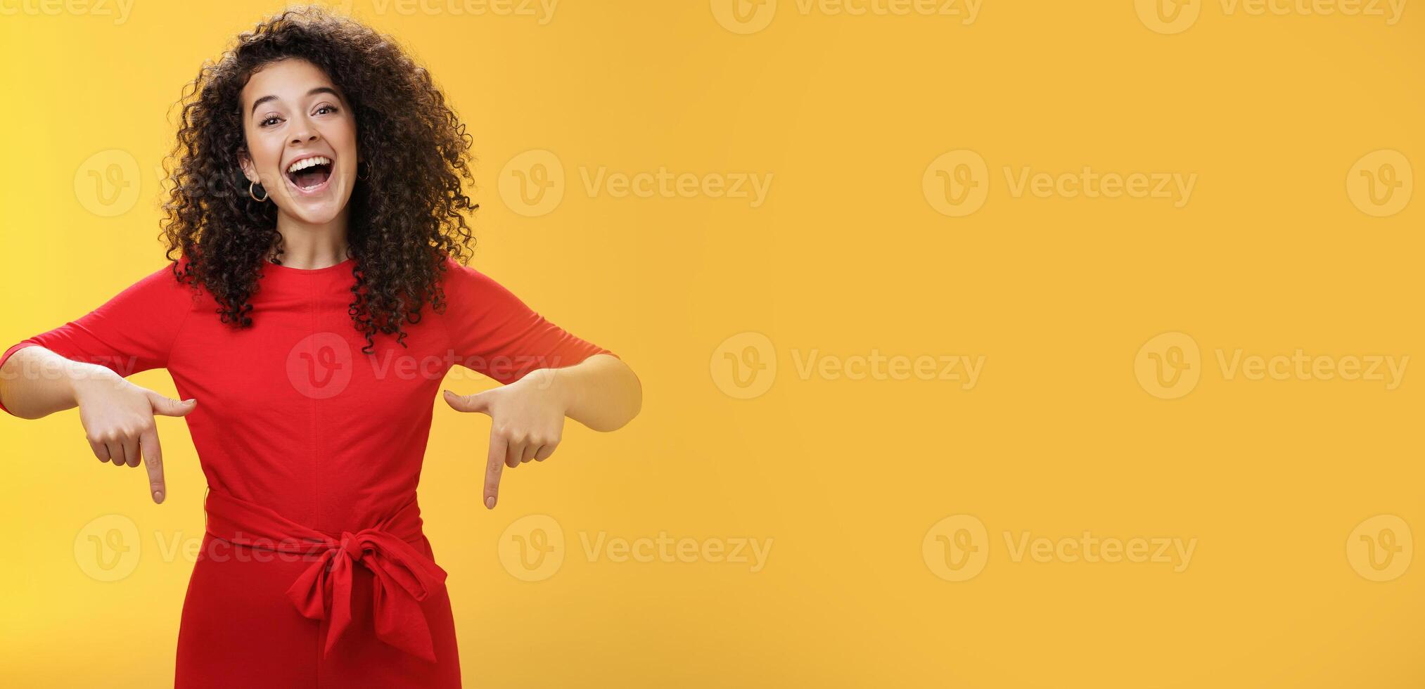 Look what I got here. Charismatic carefree happy charming woman with curly hair in red dress laughing with broad smile pointing down as showing awesome copy space to customers over yellow wall photo