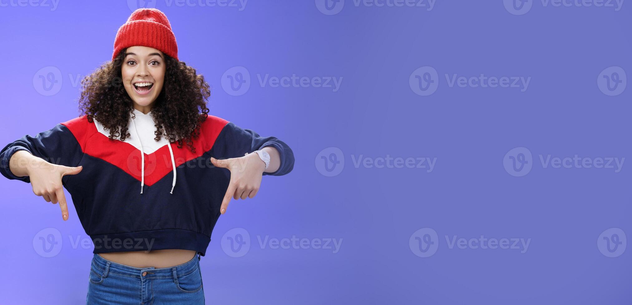 Hurry up and chek it out. Portrait of charismatic joyful and excited young cute curly-haired girl in beanie and stylish outfit pointing down, smiling broadly inviting join fun over blue background photo