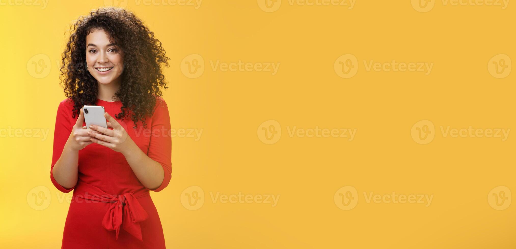 Girl messaging friend to tell all hot rumors holding mobile phone in hands smiling broadly and excited at camera as using smartphone browsing in net or edditing in app over yellow background photo