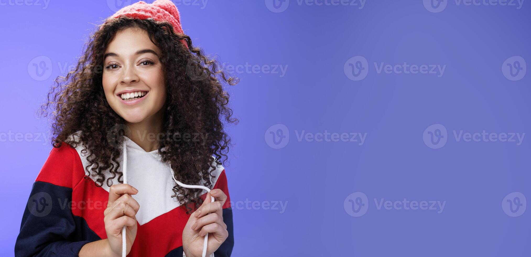 Close-up shot of carefree attractive feminine girl with curly hair in beanie playing with sweatshirt as posing over blue background, smiling at camera having fun and positive attitude photo