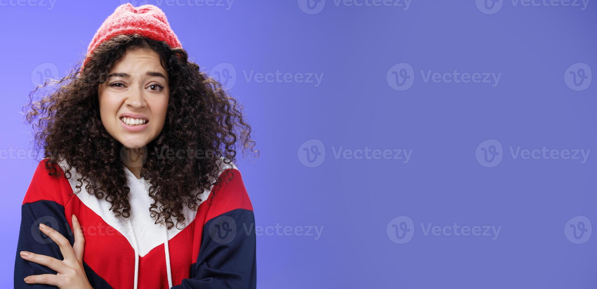 Woman with curly hair in winter beanie feeling uncomfortable and discomfort clenching teeth and frowning intense as hugging herself insecure and awkward, unwilling to say cruel rejection, feel awkward photo