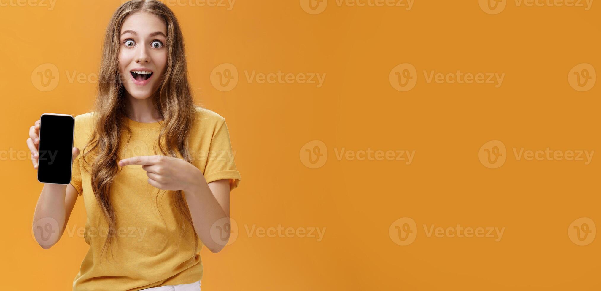 Girl surprised with awesome smartphone features. Portrait of delighted and amazed cute european slender woman with wavy natural hairstyle showing cellphone pointing at gadget screen at app photo