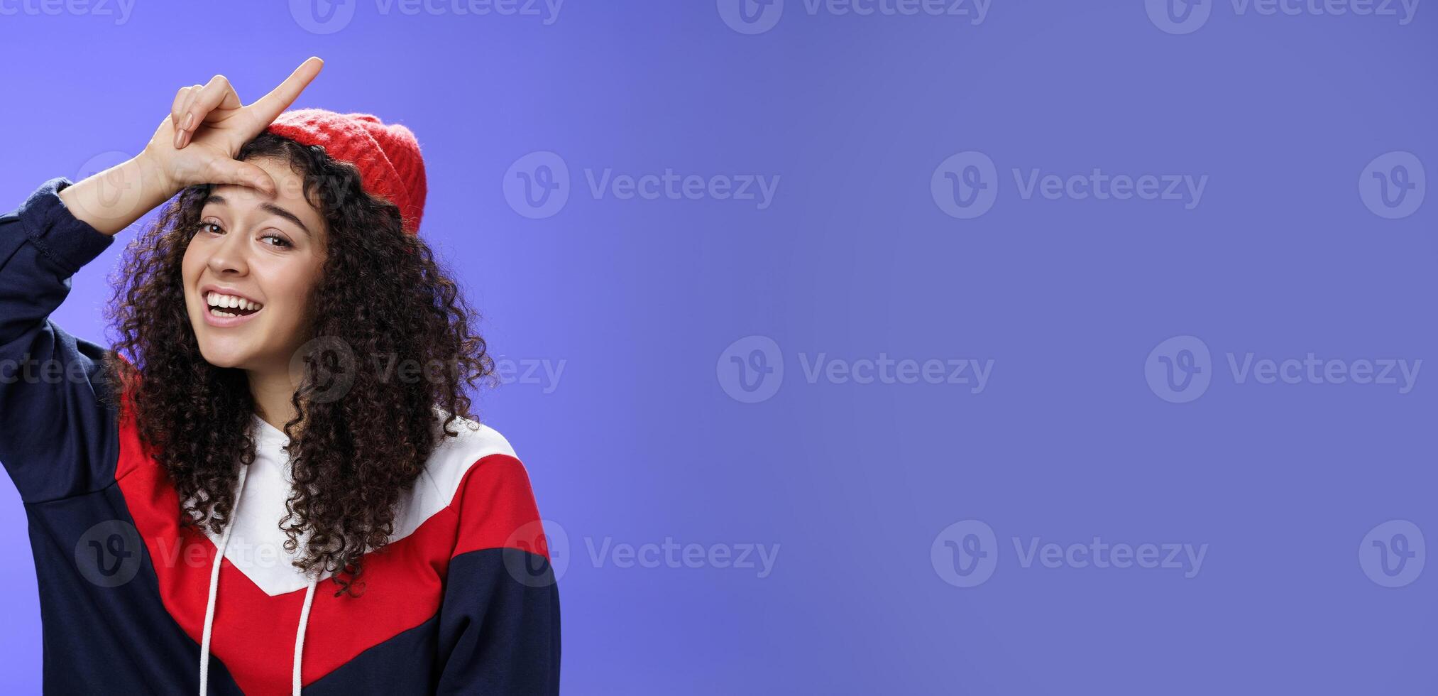 Girl triumphing and mocking friend as winning showing loser gesture on forehead and laughing having fun standing pleased and happy over blue background, smiling at camera joyfully photo