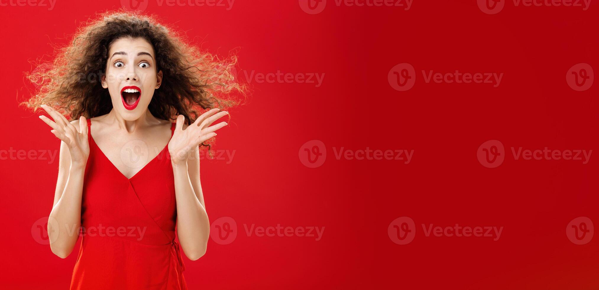 Portrait of surprised and astonished speechless curly-haired woman in red luxurious dress dropping jaw from amazement popping eyes at camera and gesturing emotive and shocked over background photo