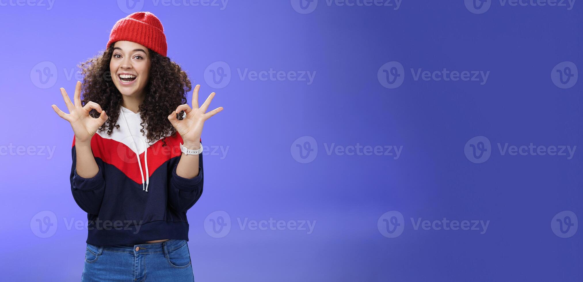 Got under control. Portrait of happy charming smiling curly-haired female in warm winter hat and sweatshirt smiling broadly and showing okay or excellent gesture as approving, liking cool movements photo