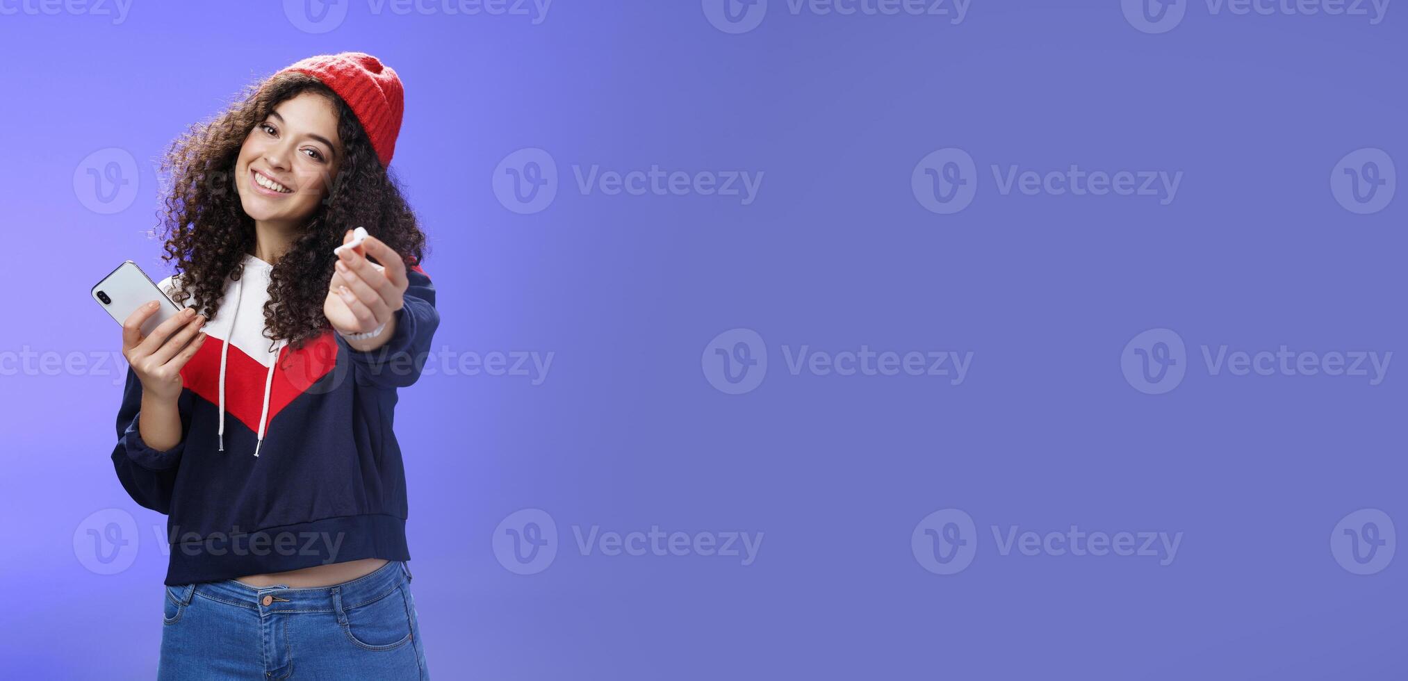Wanna listen music with me. Portrait of friendly charming and cute woman in warm beanie extending arm towards camera to suggest take wireless earphone and enjoy song together, holding smartphone photo