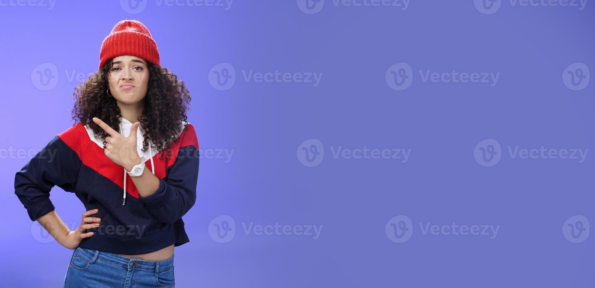 Girl feleing sad missing good chance, such shame. Portrait of displeased woman regretting frowning and pursing lips gloomy pointing at upper left corner upset wearing warm beanie over blue wall photo