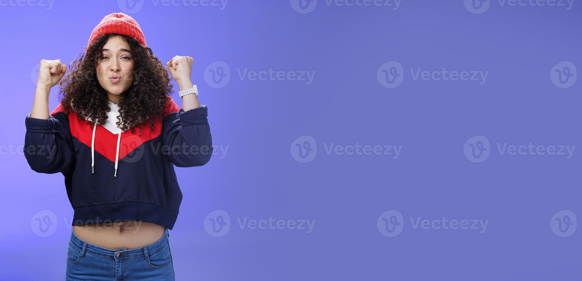 Charming young 20s girl with curly hair in warm beanie raising hands as belly showing under sweatshirt triumphing, celebrating awesome win folding lips from joy and excitement over blue background photo