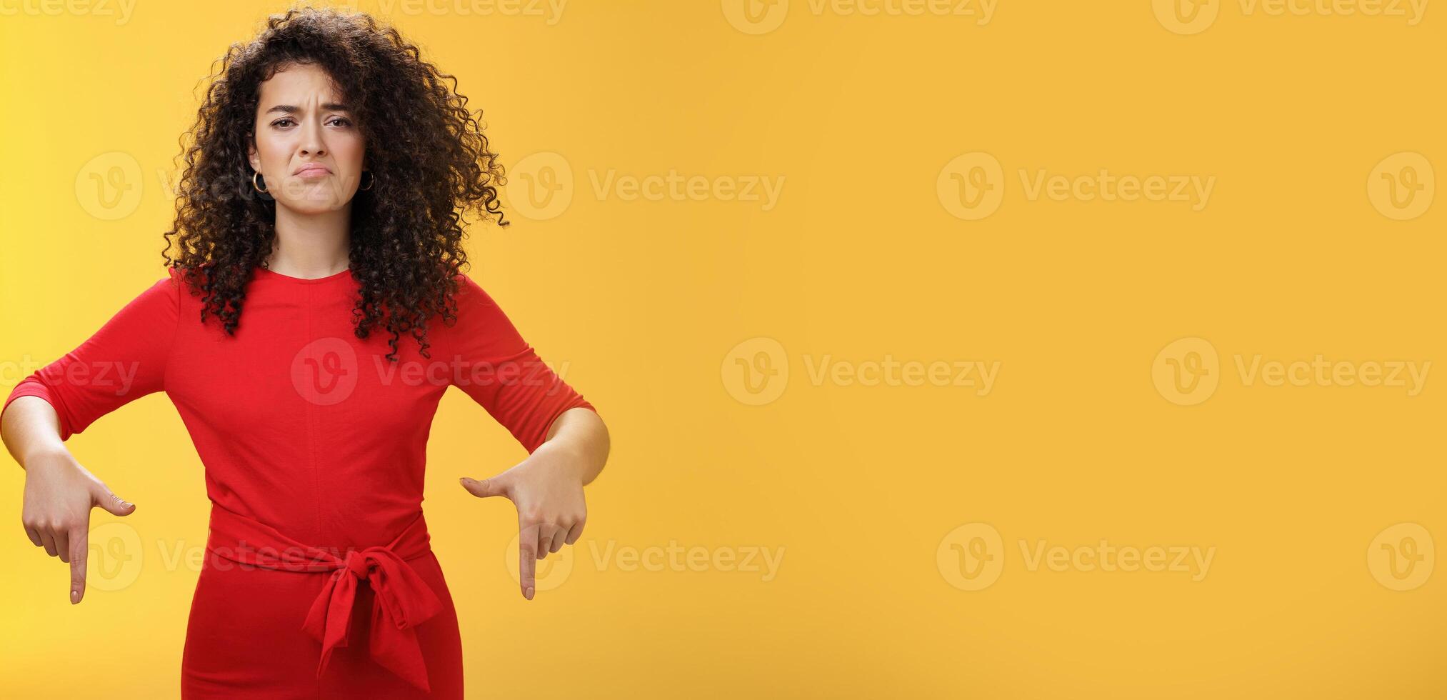 Gloomy disappointed cute silly curly-haired female in red dress frowning upset and making sad smile as pointing down with regret and unsatisfied expression, jealous or displeased over yellow wall photo