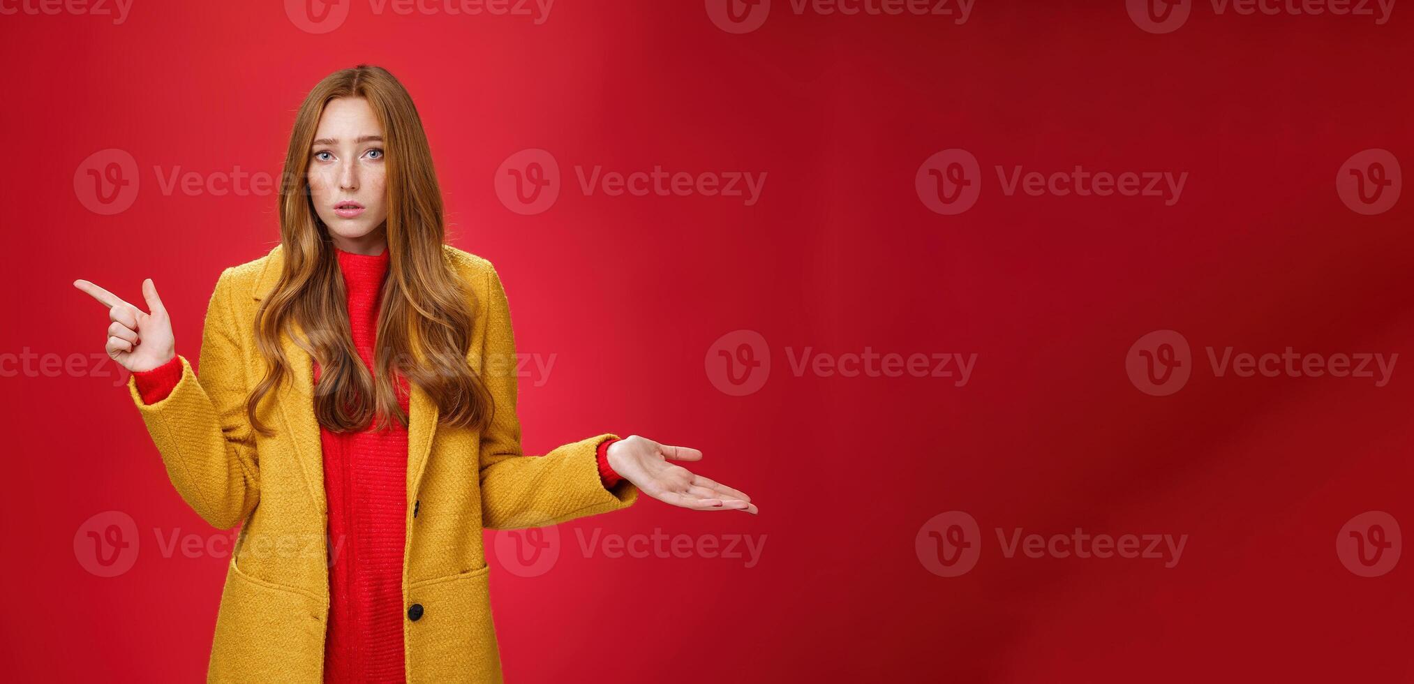 Frustrated and confused young cute female with ginger hair in yellow coat shrugging as pointing questioned left waiting explanation as asking question clueless and puzzled over red background photo