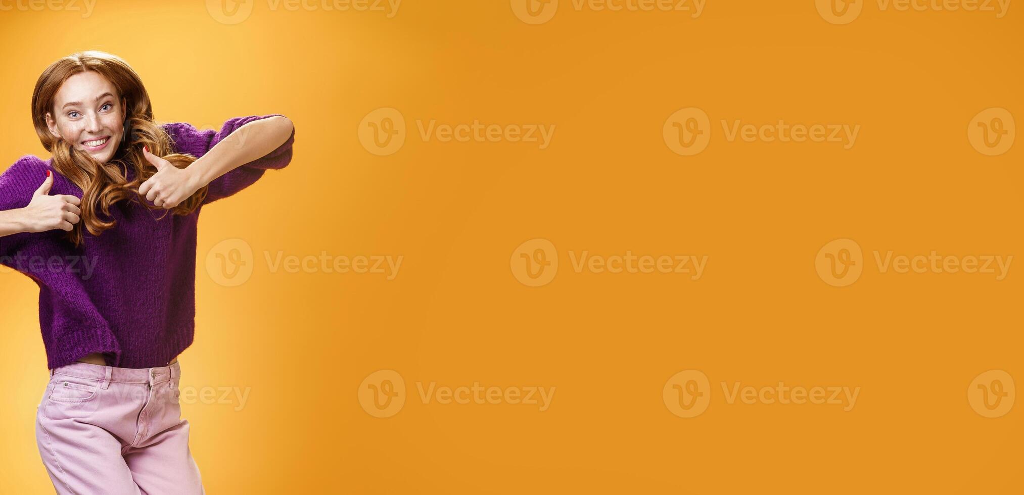 Optimistic and joyful bright ginger girl jumping happily and cheerful having fun showing thumbs up in like and approval giving positive feedback of super cool idea or concept over orange wall photo