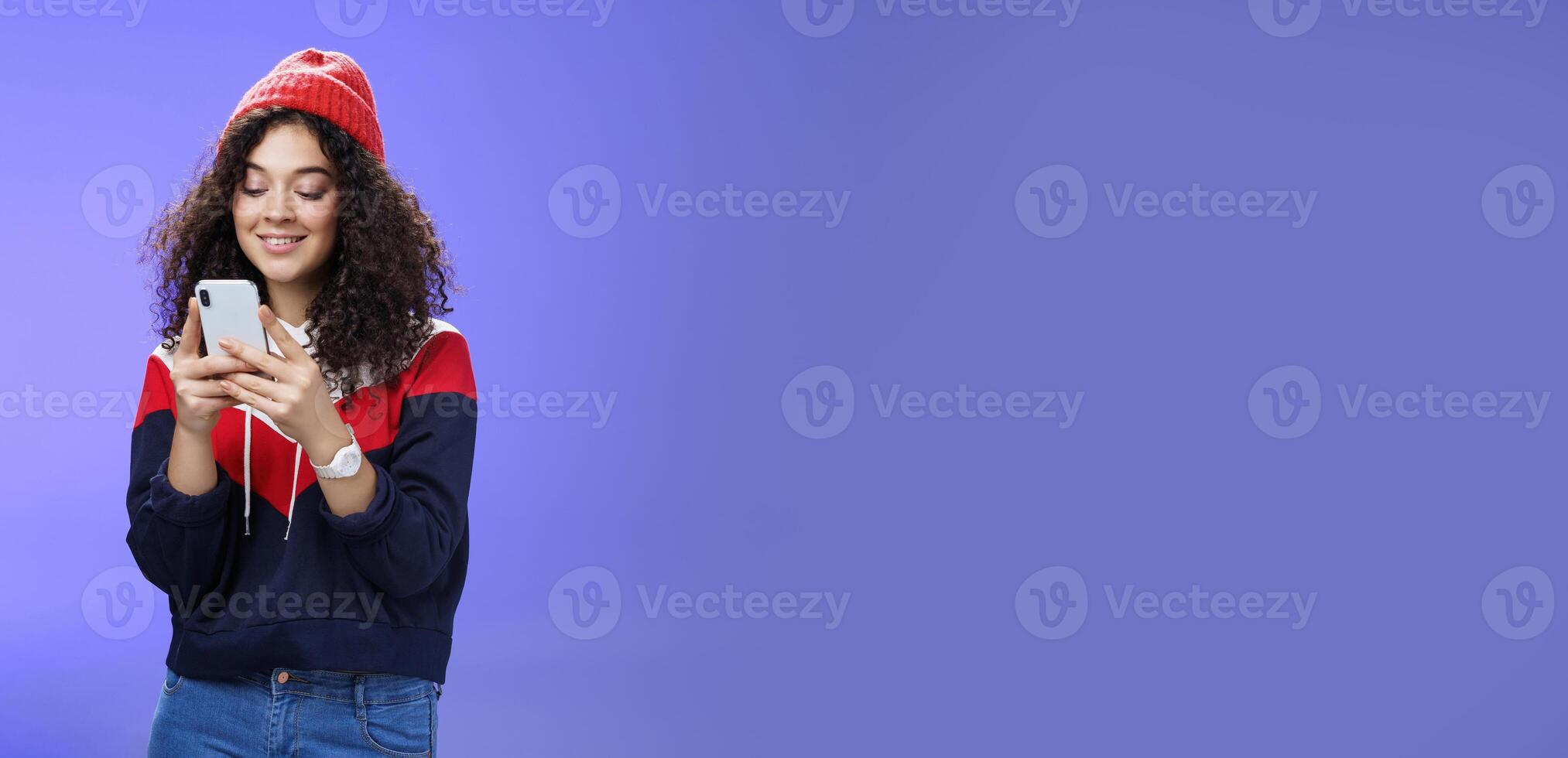 Woman searching in internet funny memes, holding mobile phone in hands smiling joyfully and cute at smartphone screen, typing message or browsing internet over blue background, wearing warm winter hat photo