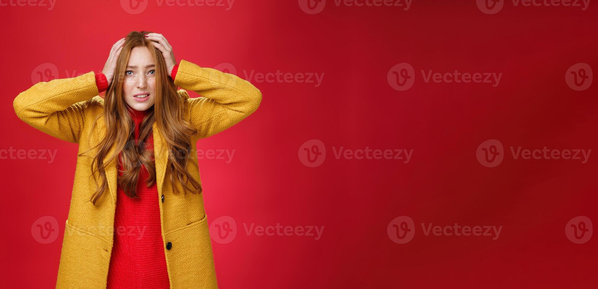 Worried and freaked out redhead girl holding hands on head grimacing troubled and concerned clenching teeth, frowning worrying and feeling nervous for project going fail over red background photo