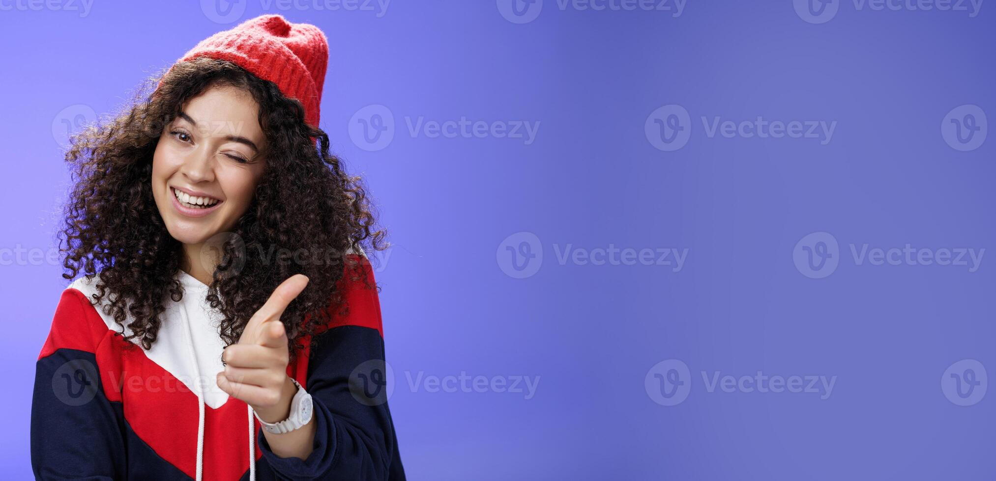 Friendly-looking sociable and stylish woman with curly hair in red beanie winking joyfully and pointing with finger pistol at camera as greeting mate being cool and confident over blue wall photo
