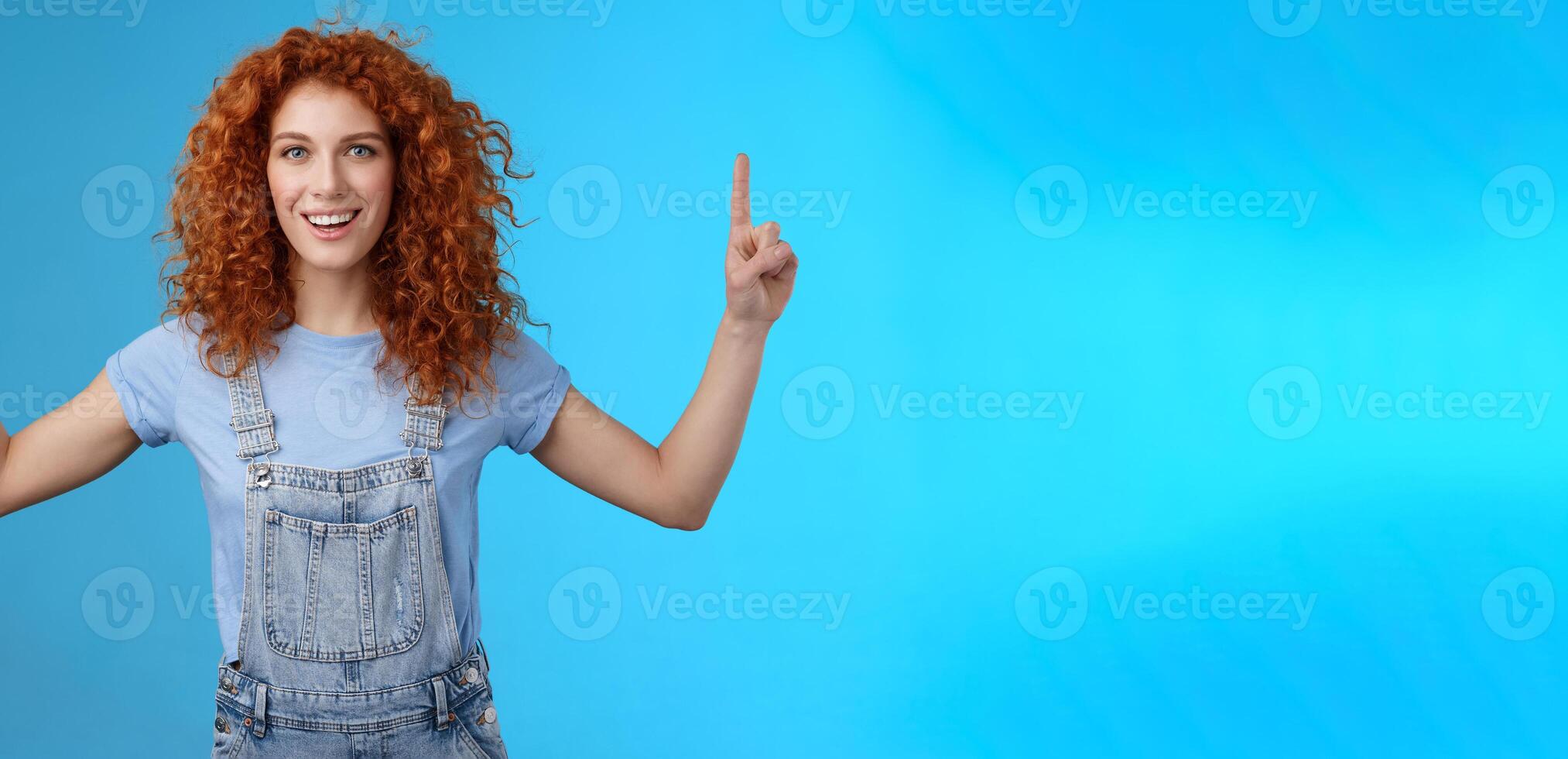 Energized enthusiastic good-looking redhead cheerful curly girl pointing up index fingers motivated present awesome copy space smiling delighted glad summer warm days near beach blue background photo