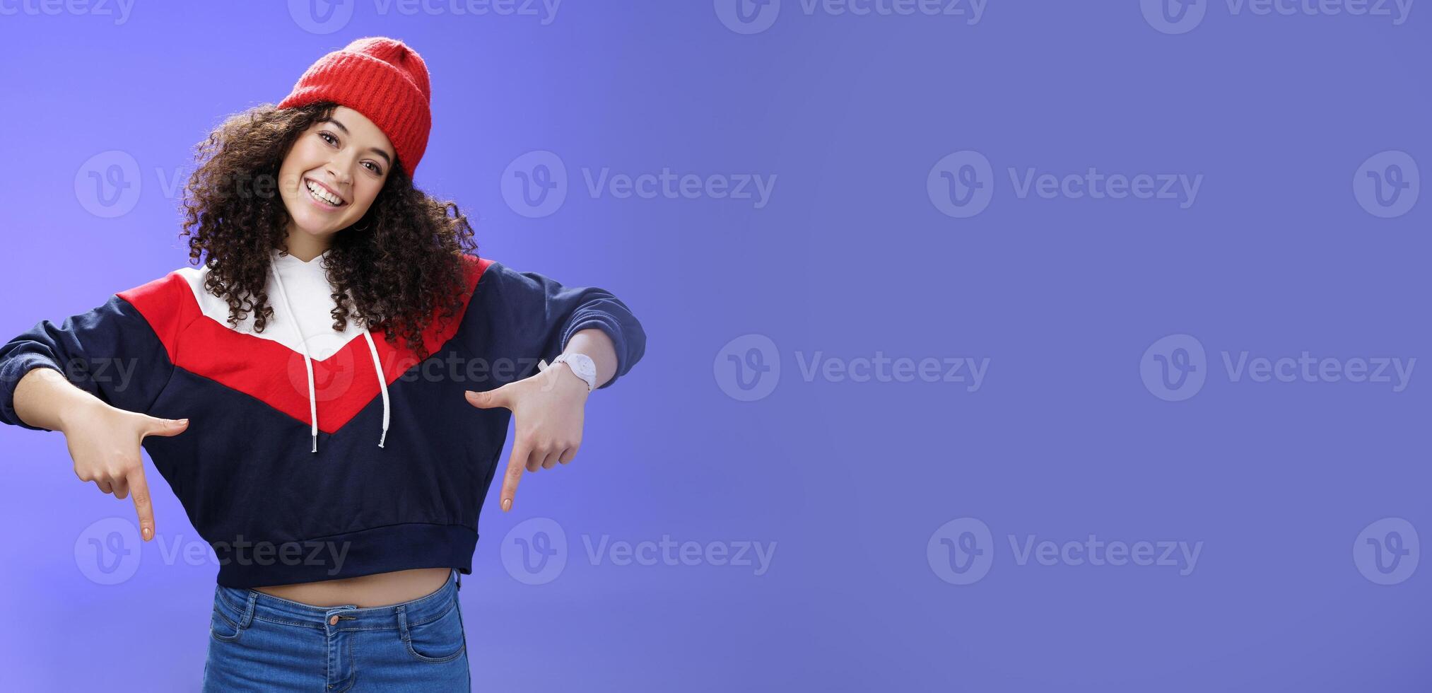 Waist-up shot of cool and stylish young woman with curly hair smiling flirty and joyful pointing down showing promotion as tilting head happily and posing over blue background in outdoor clothes photo