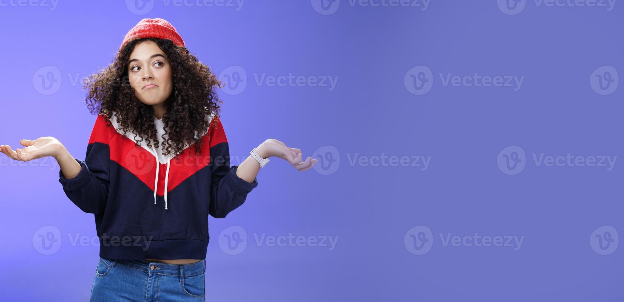Not getting clue. Cute silly woman with curly hair shrugging with pursed lips and unbothered expression holding hands sideways looking right being unaware and confused, uncertain what answer photo