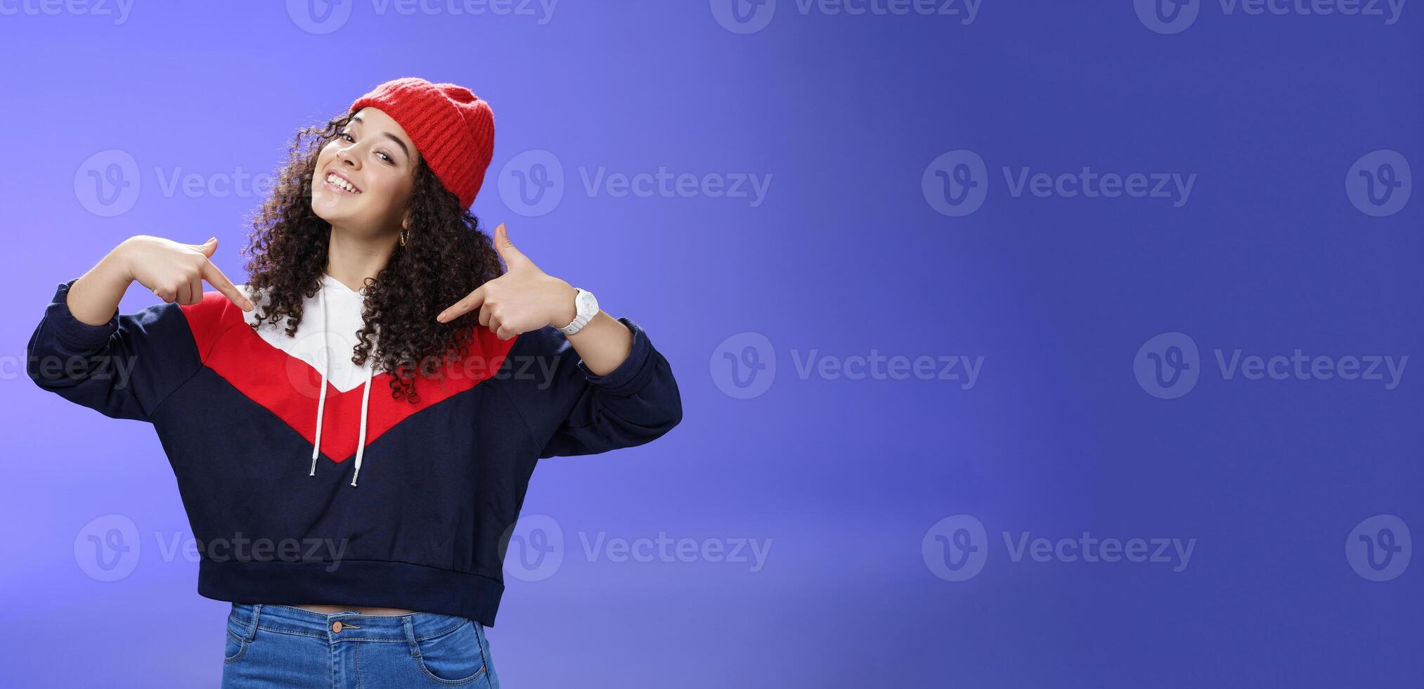 Proud and self-satisfied happy young energized stylish female model in warm beanie brag about own achievements as pointing at herself raising head and smiling produly posing delighted over blue wall photo