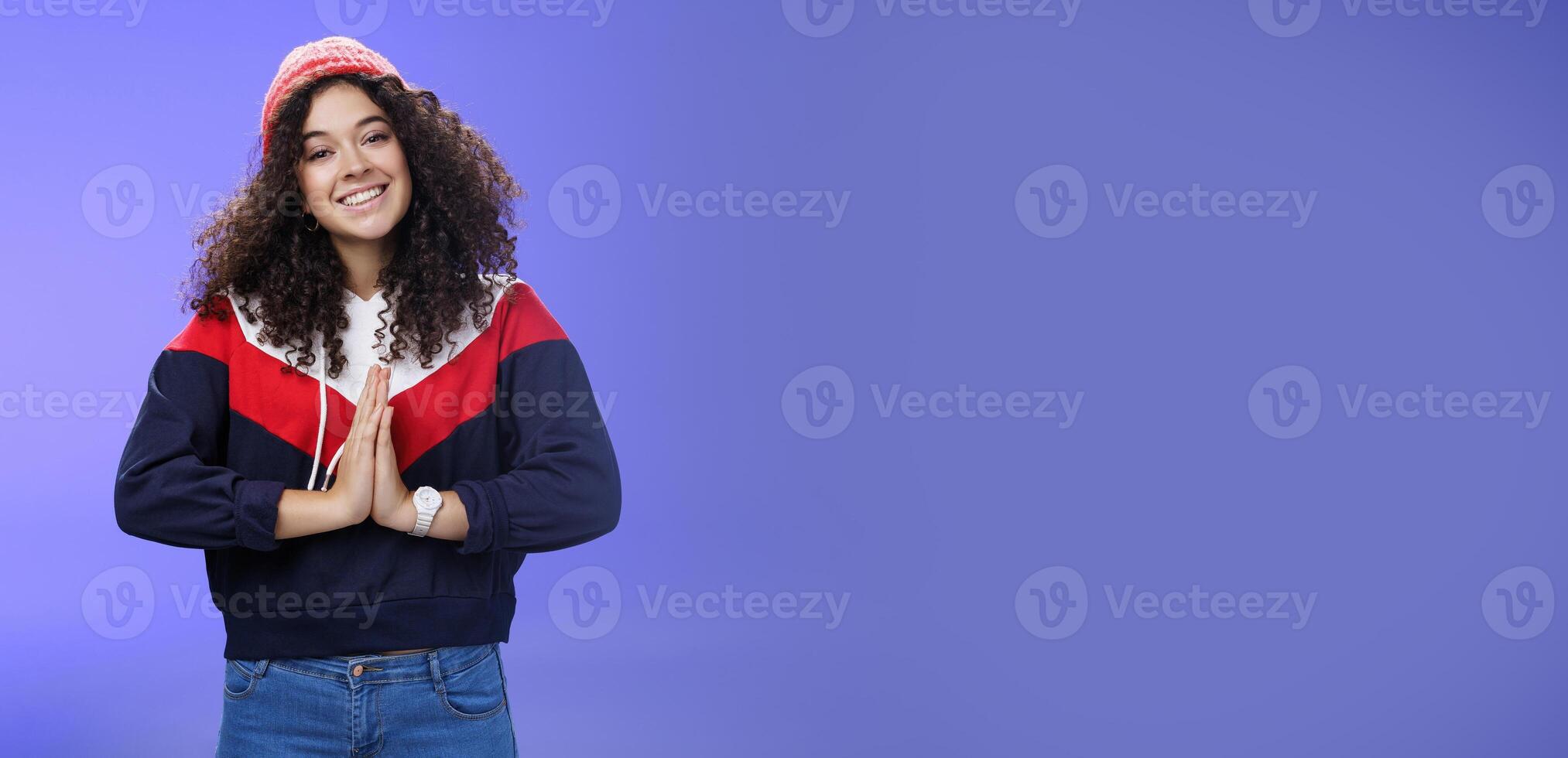 Portrait of charming curly-haired feminine girlfriend begging for favour as posing over blue background in winter outfit with hat, holding hands in pray smiling with angel look and friendly gaze photo