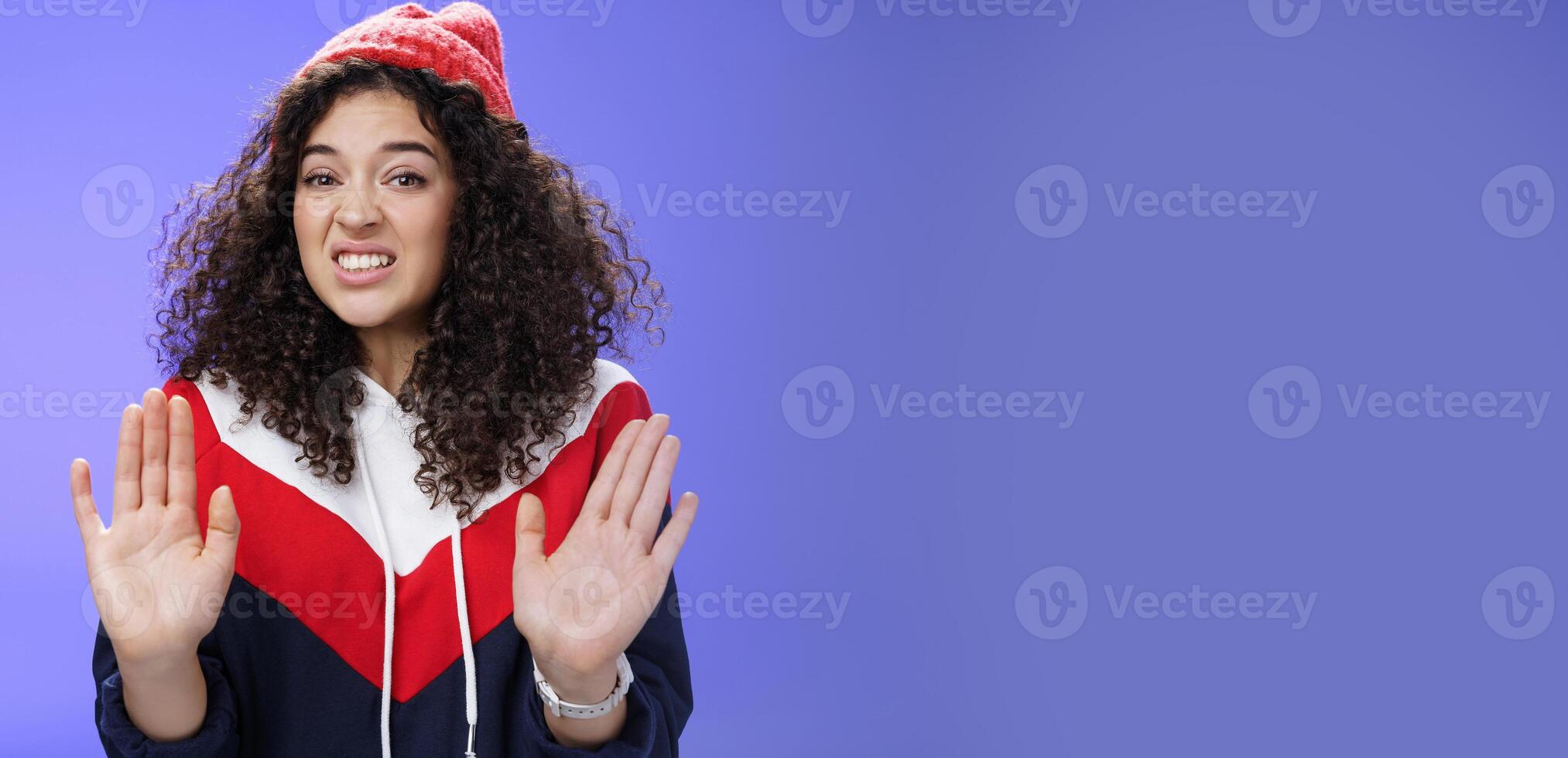 Displeased european woman with curly hair raising palms near chest and waving in refusal and rejection gesture grimacing unsatisfied and unwilling to participate in suspicious action over blue wall photo
