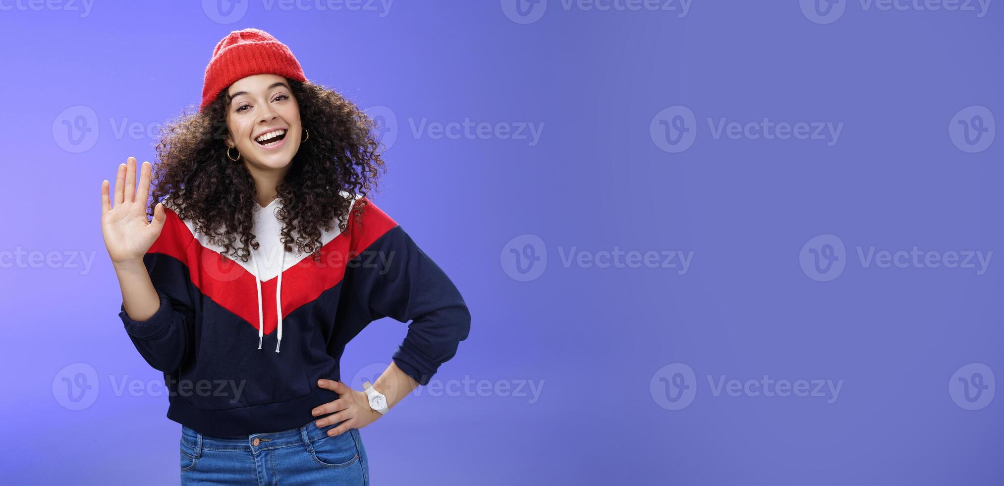 Hey nice to meet you. Charming female ski couch in cute red beanie with curly hair waving hello with palm and smiling broadly greeting newbies as teaching winter sports over blue background photo