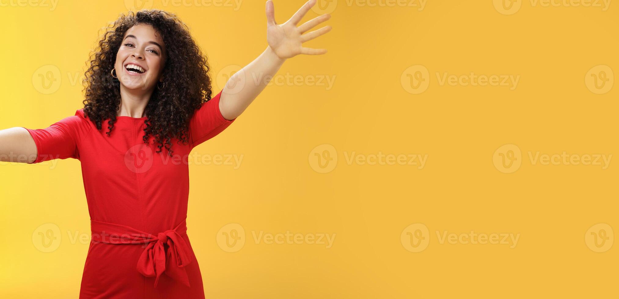 Come into my arms. Portrait of friendly and loving, caring charming woman with curly hair in red casual dress spread hands as wanting give hug smiling broadly at camera giving warm welcome or cuddle photo
