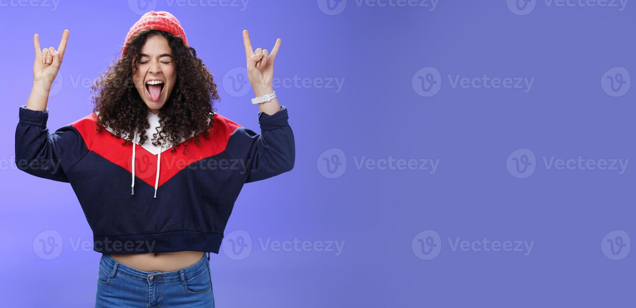 Waist-up shot of rebellious and carefree woman having fun feeling excited and awesome at party close eyes sticking out tongue and showing rock-n-roll signs, wearing beanie and warm clothes photo