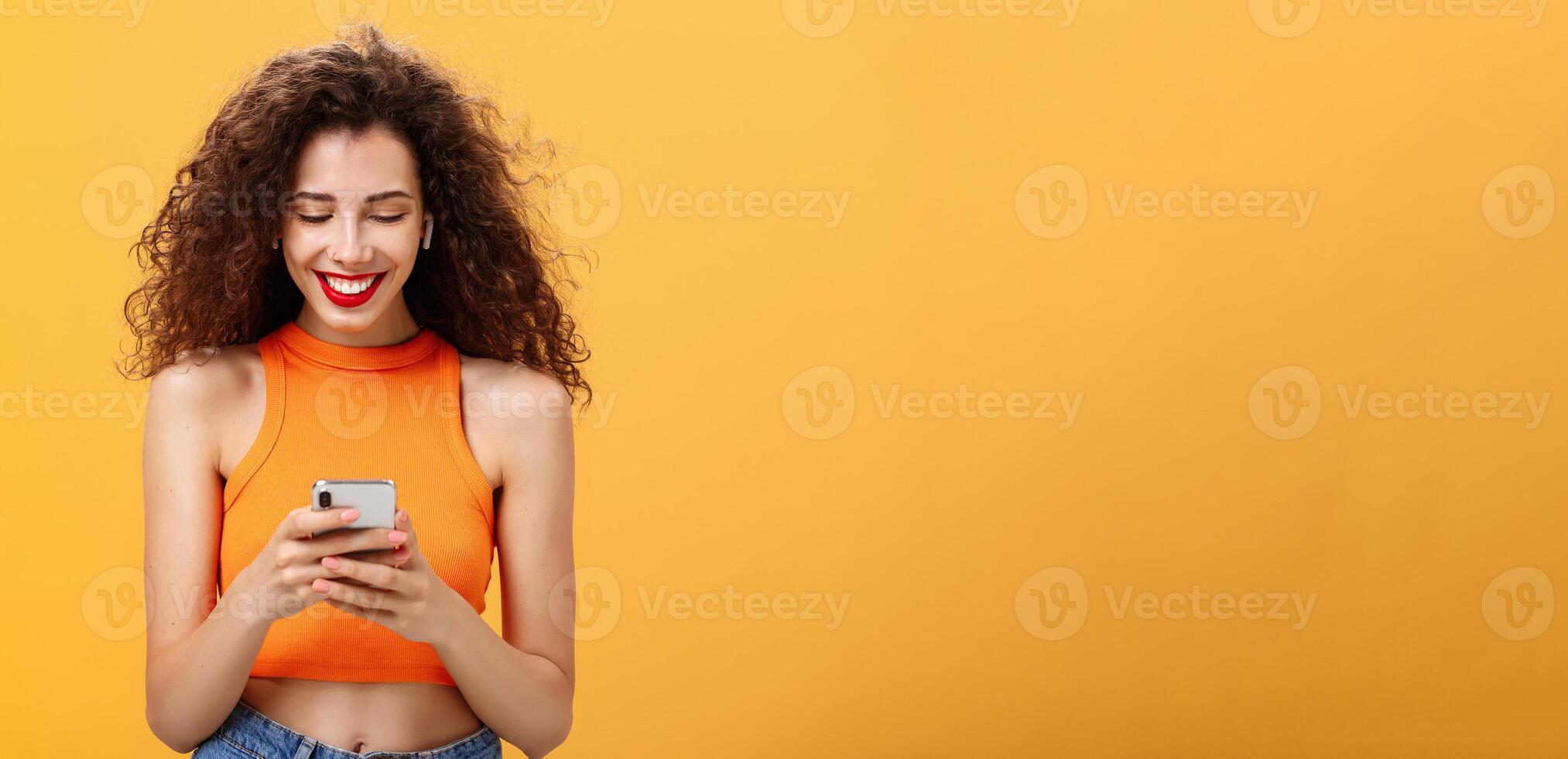 Stylish young attractive woman with curly hairstyle, nose ring and red lipstick picking song, typing in smartphone listening music in wireless earphones smiling at device screen over orange wall photo