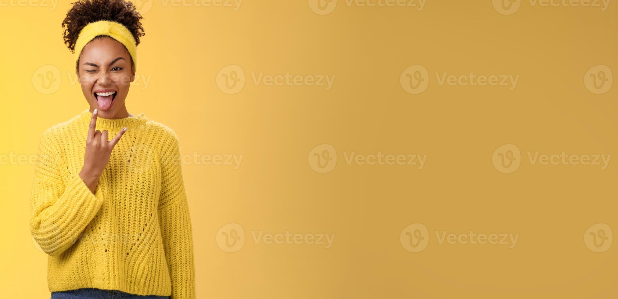Excited charming smiling daring sassy young african american woman having fun show tongue rock-n-roll heavy metal gesture winking happily enjoying cool awesome party, standing yellow background photo