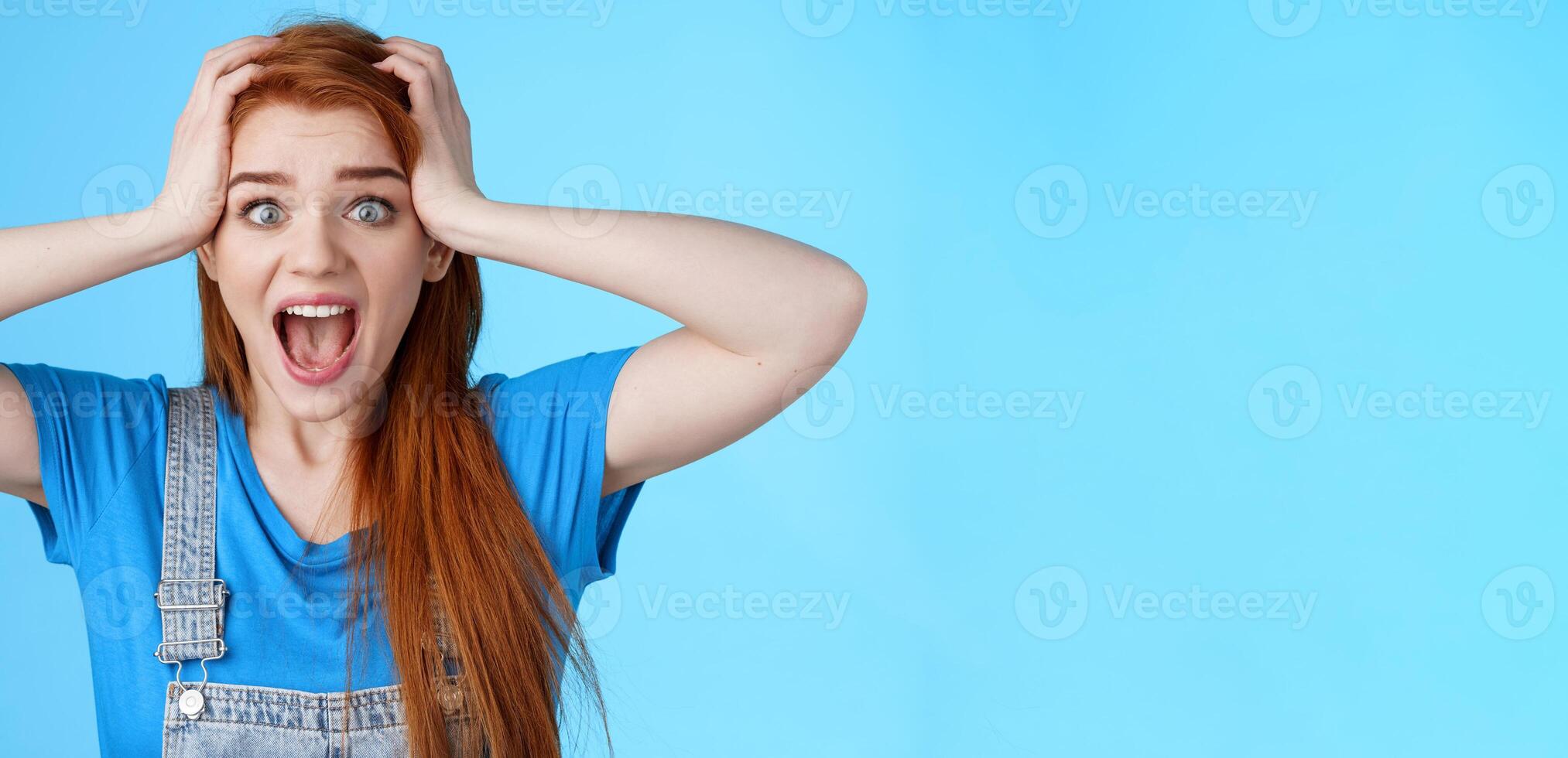 Shocked redhead girl shouting panic, feel pressured deadlines, scared see mess, grab head, scream frightened distressed, stare camera hopeless, anxiously react losing lottery, blue background photo