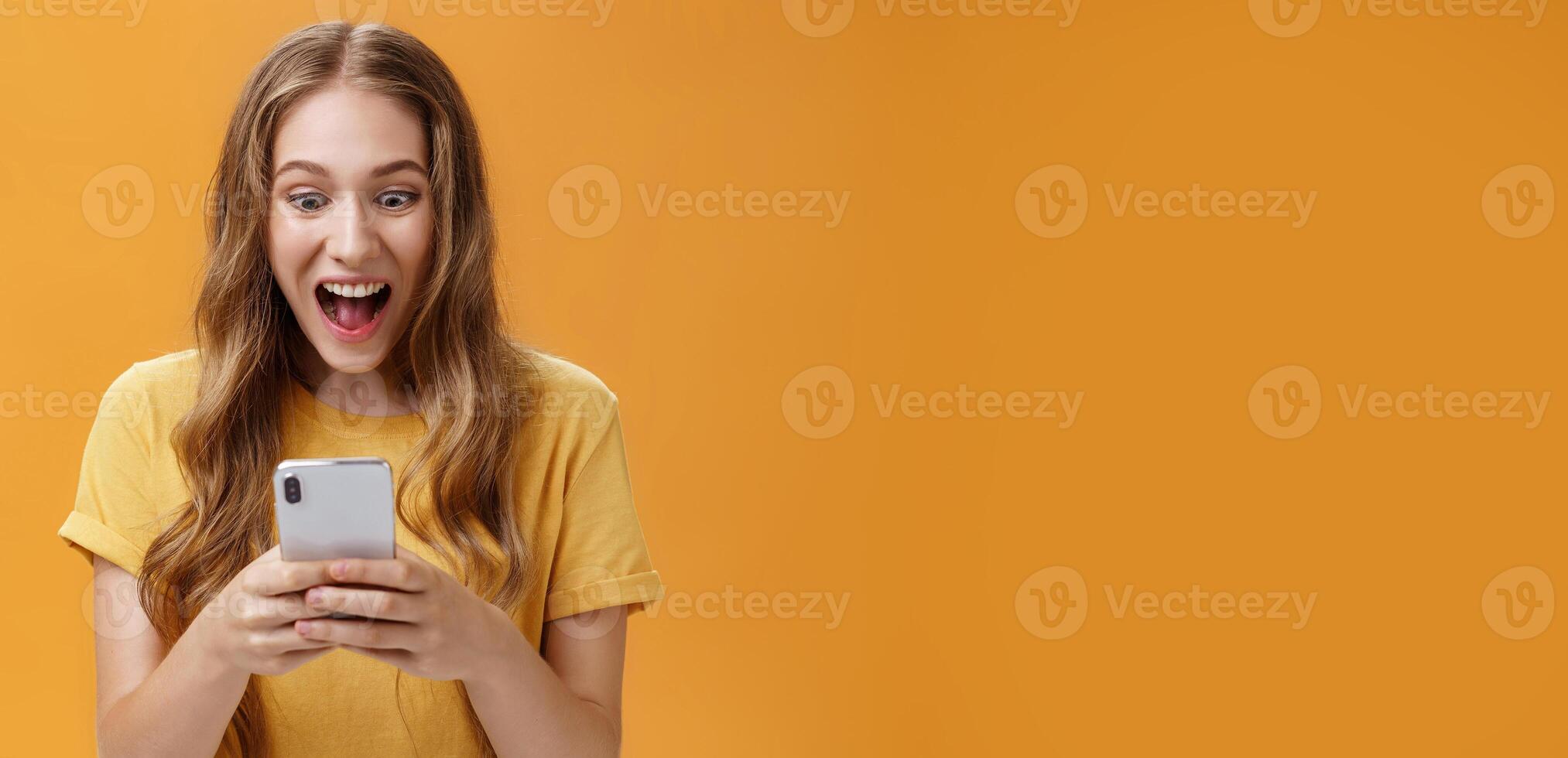Excited and thrilled charismatic young happy girl in yellow t-shirt smiling with opened from joy mouth holding smartphone staring at cellphone screen astonished carried away with cool phone game photo