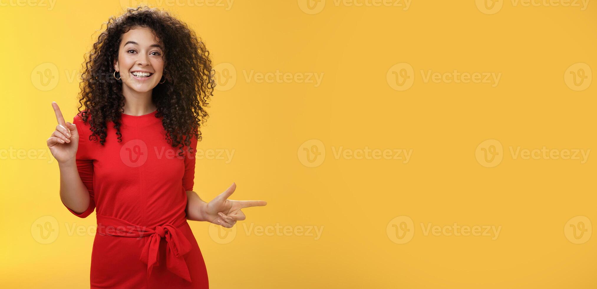 Charismatic cute and happy young curly-haired european female in red dress dancing and pointing up and right, showing sideways as presenting choices picking, making decision and smiling at camera photo
