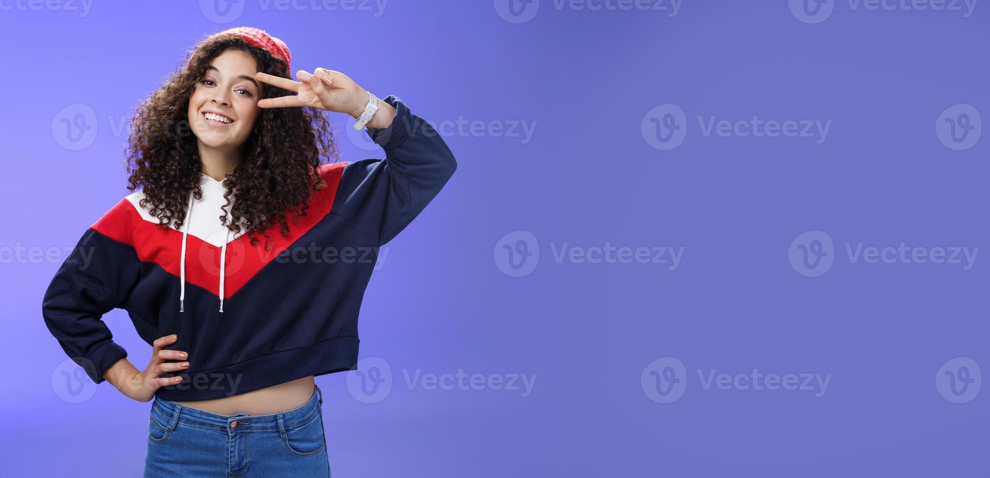 Attractive feminine woman with curly hairstyle in hat and sweatshirt showing peace or victory sign around eye and smiling carefree having fun playing in yard with snow over blue background photo