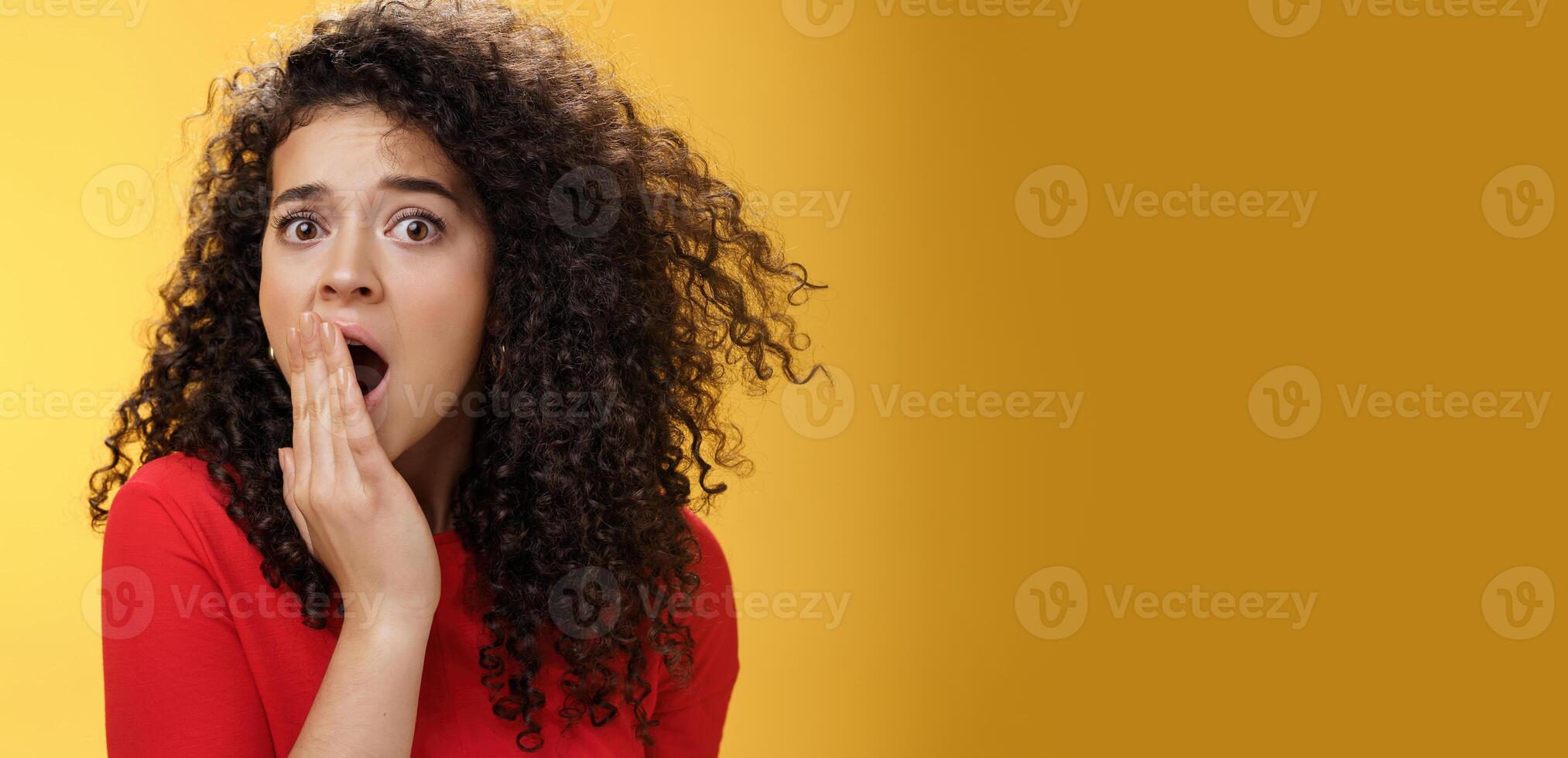Close-up shot of concerned and shocked worried woman with curly hairstyle open mouth wide and covering it with palm popping eyes at camera impressed and upset over yellow background photo