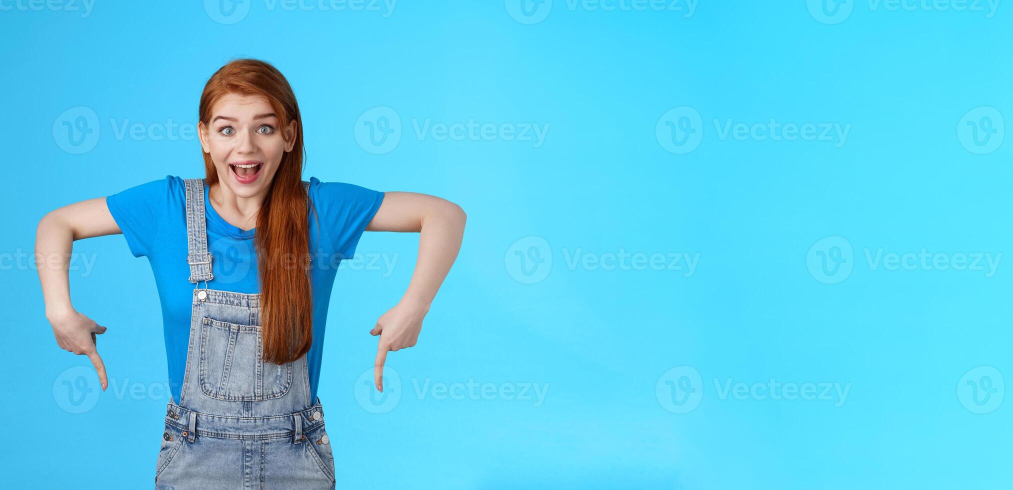 Amused wondered excited cute 20s european woman, pointing down, bottom copy space, look amazed admiration camera, describe amazing product, recommend site, stand blue background photo
