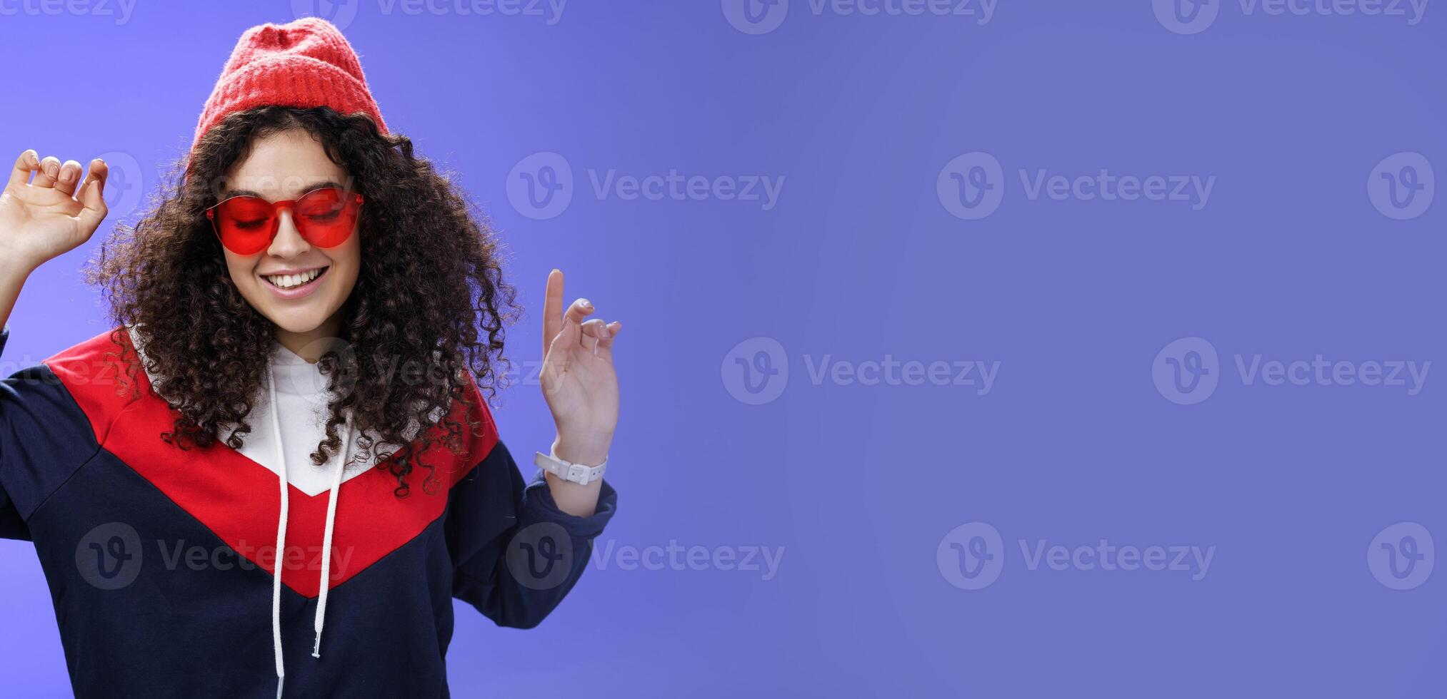 Girl feeling awesome having fun and enoying cool party, dancing with raised hands looking down smiling broadly wearing sunglasses and stylish beanie posing against blue background photo