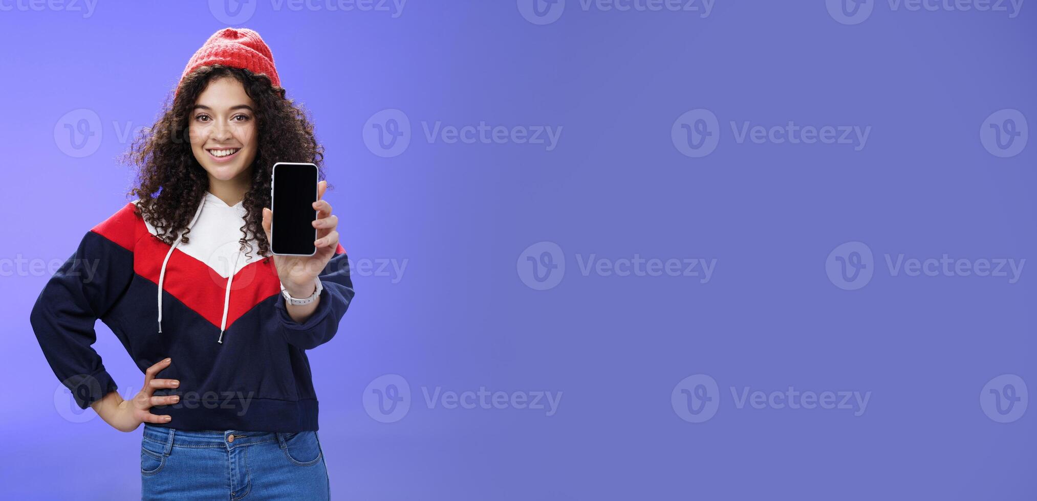 Girl showing her post as pulling smartphone screen towards camera smiling delighted waiting opinion, holding hand on waist in confident pose wearing warm winter red beanie and sweatshirt photo