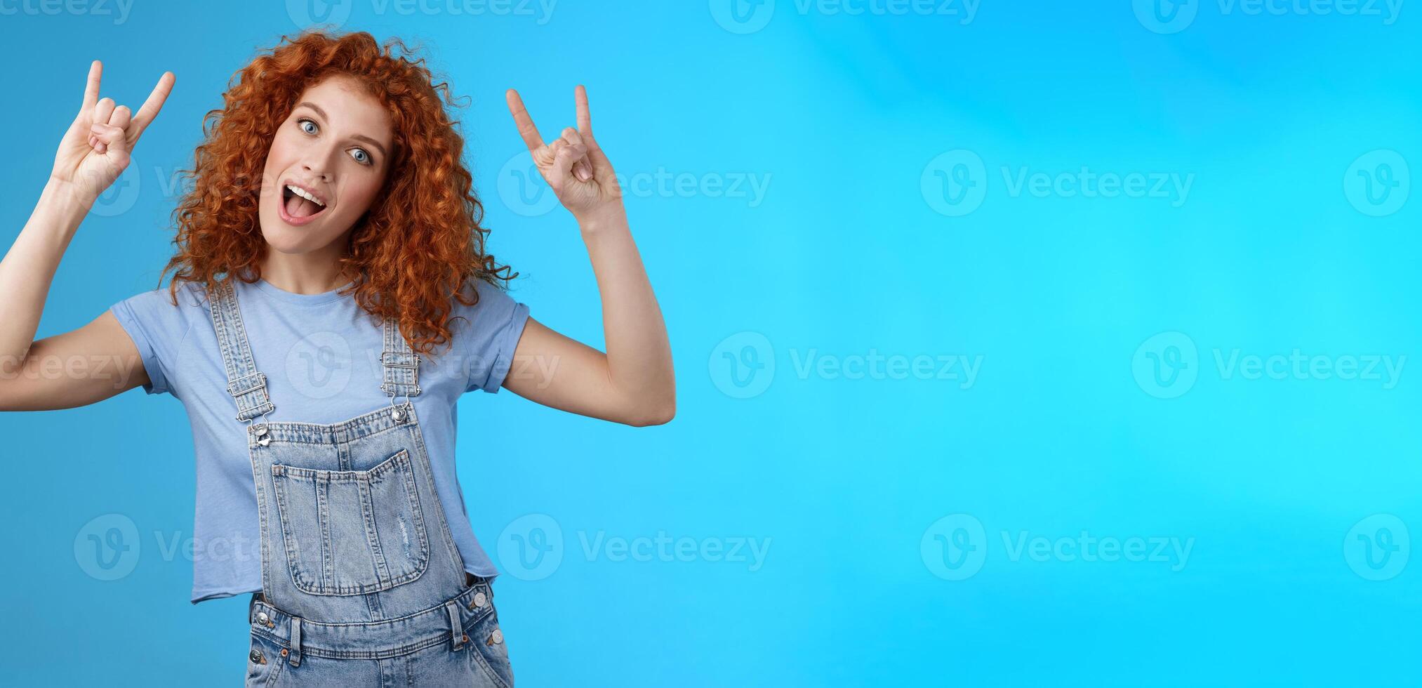 Daring cool stylish awesome redhead cheerful curly-haired girl tilt head show tongue joyfully stare camera playful raise hands rock-n-roll heavy metal gesture having fun blue background photo