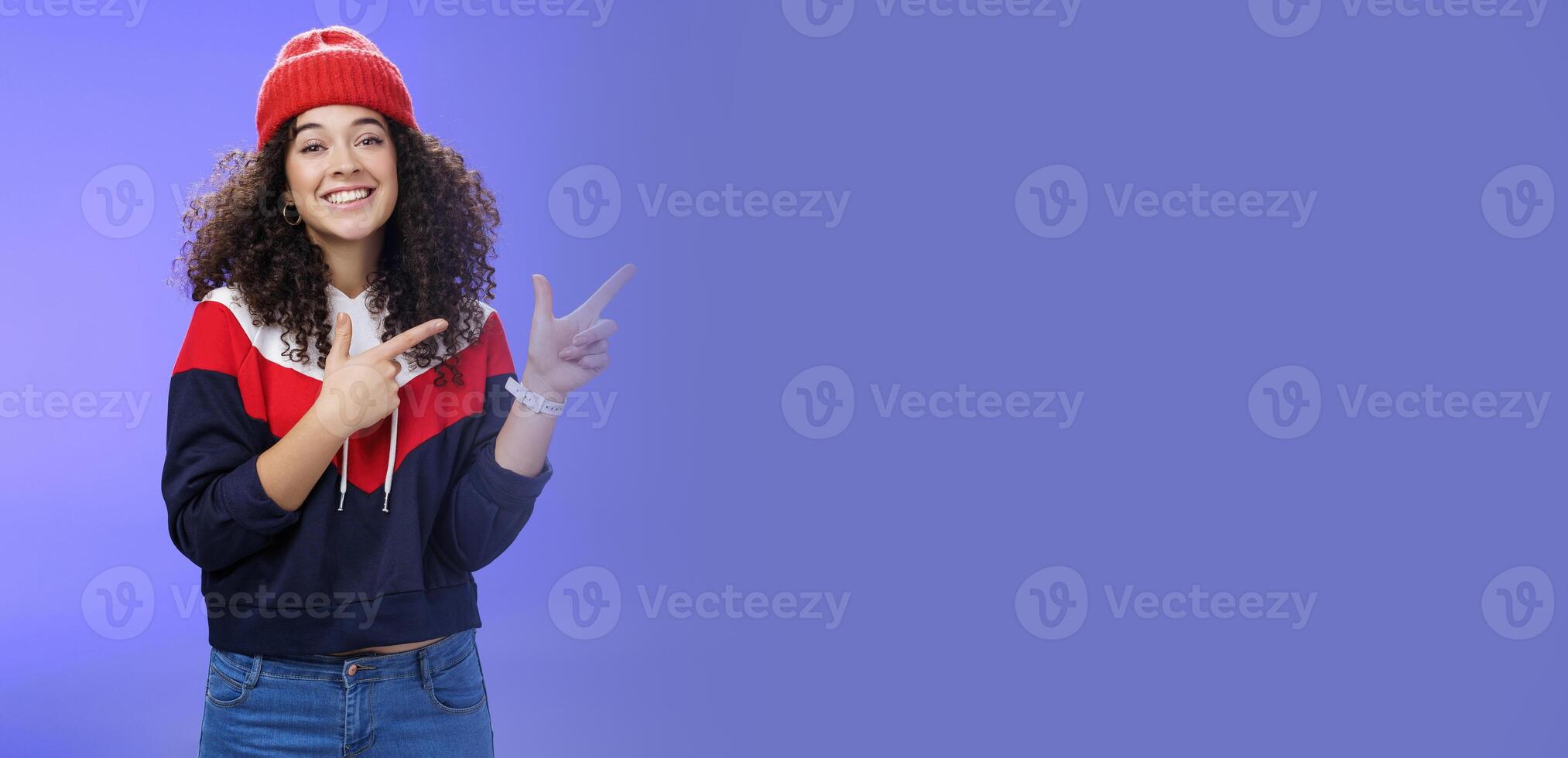 Cute female friend pointing at upper right corner with finger pistols as smiling broadly at camera wearing warm winter hat and sweatshirt skiing over blue background joyfully photo