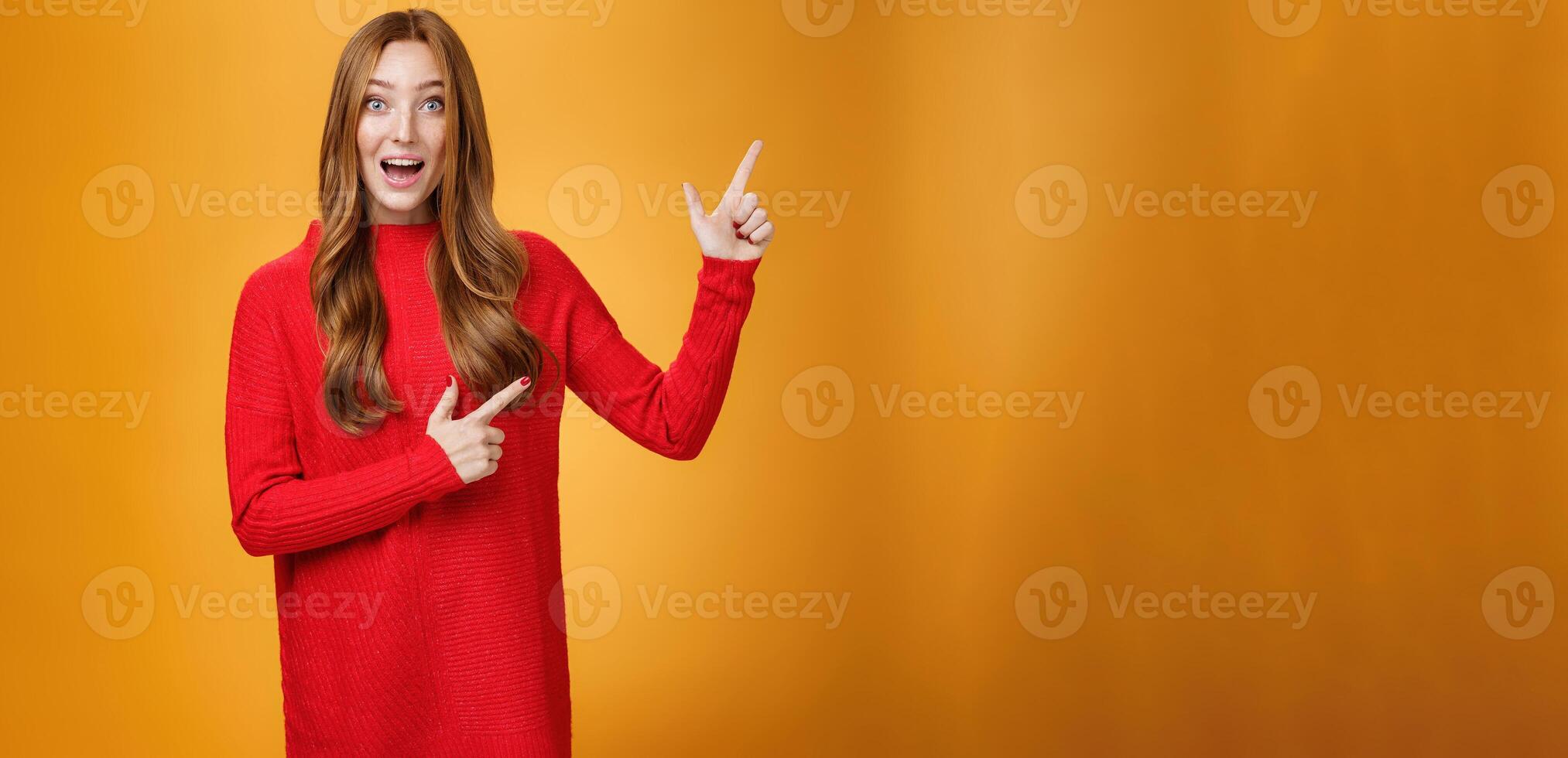 Enthusiastic and ambitious cute redhead female with freckles in red dress pointing at upper right corner smiling amazed and curious as exploring awesome new space posing over orange background photo
