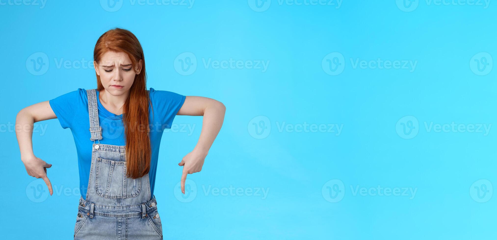 Moody upset cute redhead woman feel uneasy, frowning look pointing up jealousy and regret, cannot afford awesome new product, express sadness and sorrow, stand blue background gloomy photo